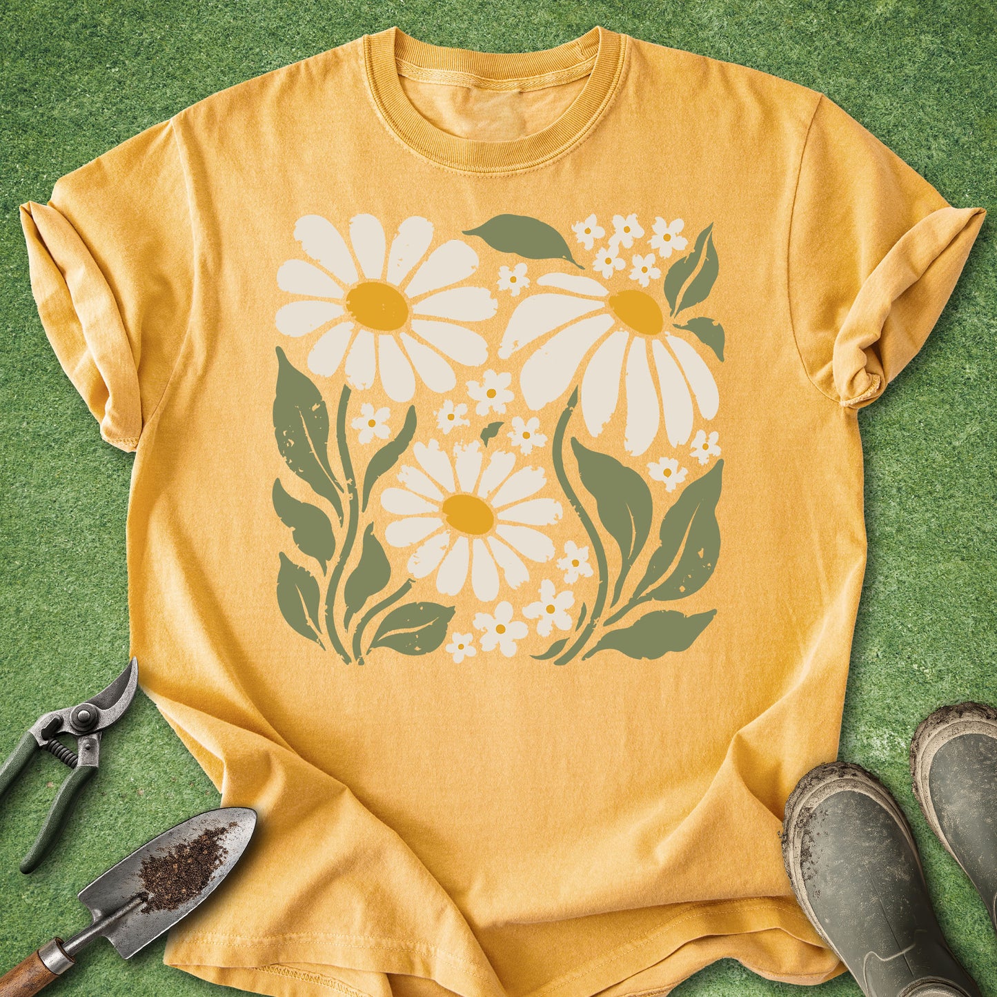 A mustard yellow short-sleeve t-shirt with a retro-style daisy and flower graphic, laid flat on green grass with gardening tools and boots nearby.