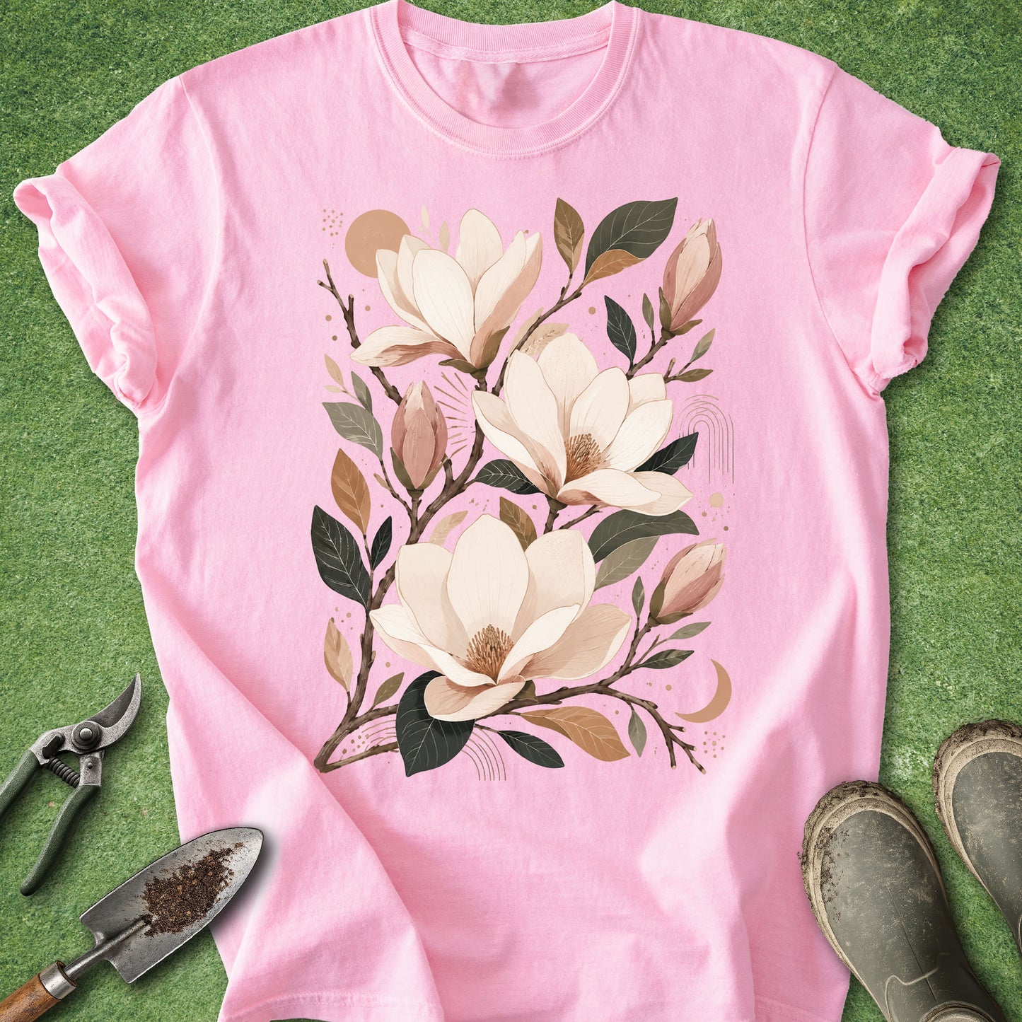 A pink short-sleeve t-shirt with a large graphic of magnolia flowers and leaves, laid flat on green grass with gardening tools and boots nearby.