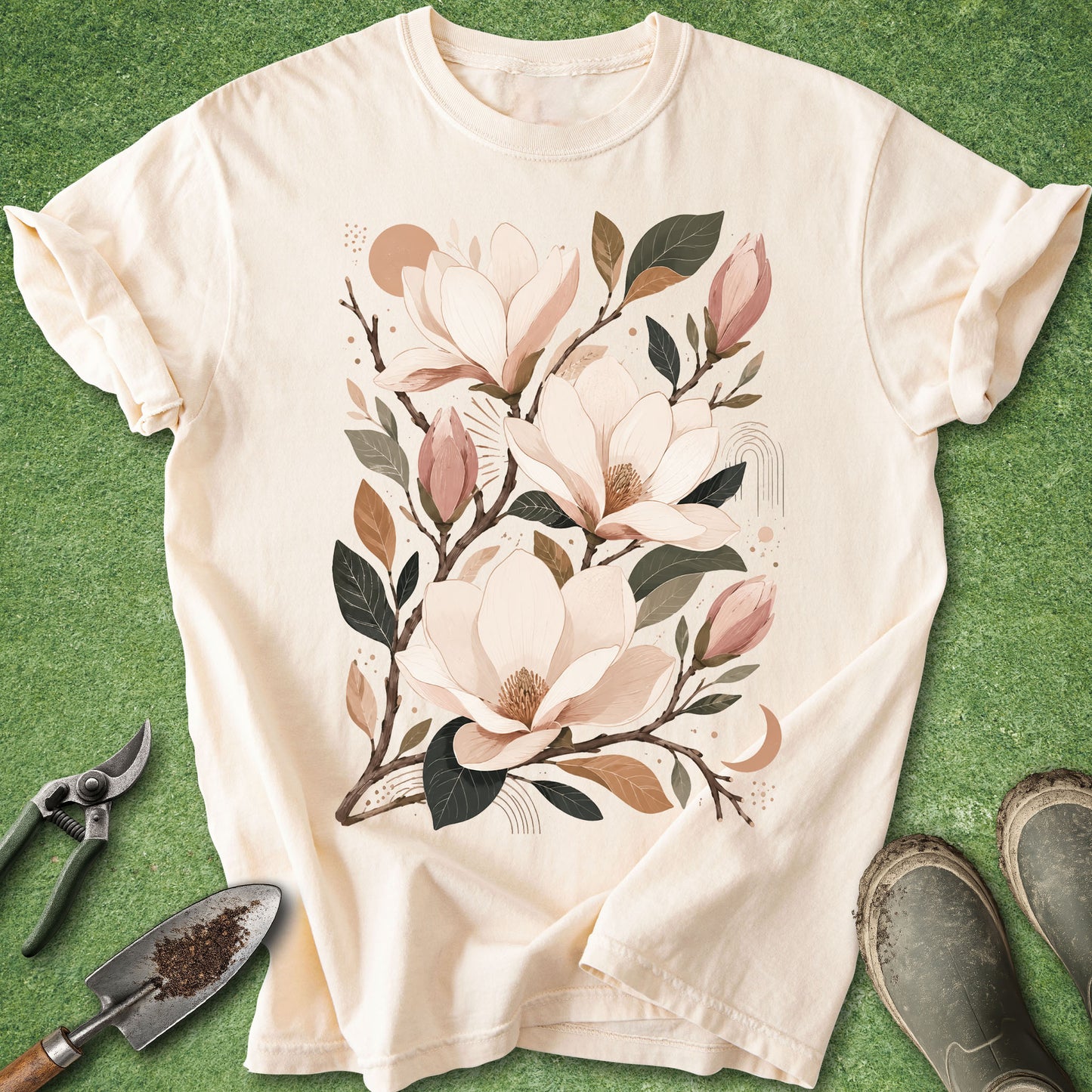 A cream-colored short-sleeve t-shirt with a large graphic of magnolia flowers and leaves, laid flat on a green grass-like surface with gardening tools and boots nearby.