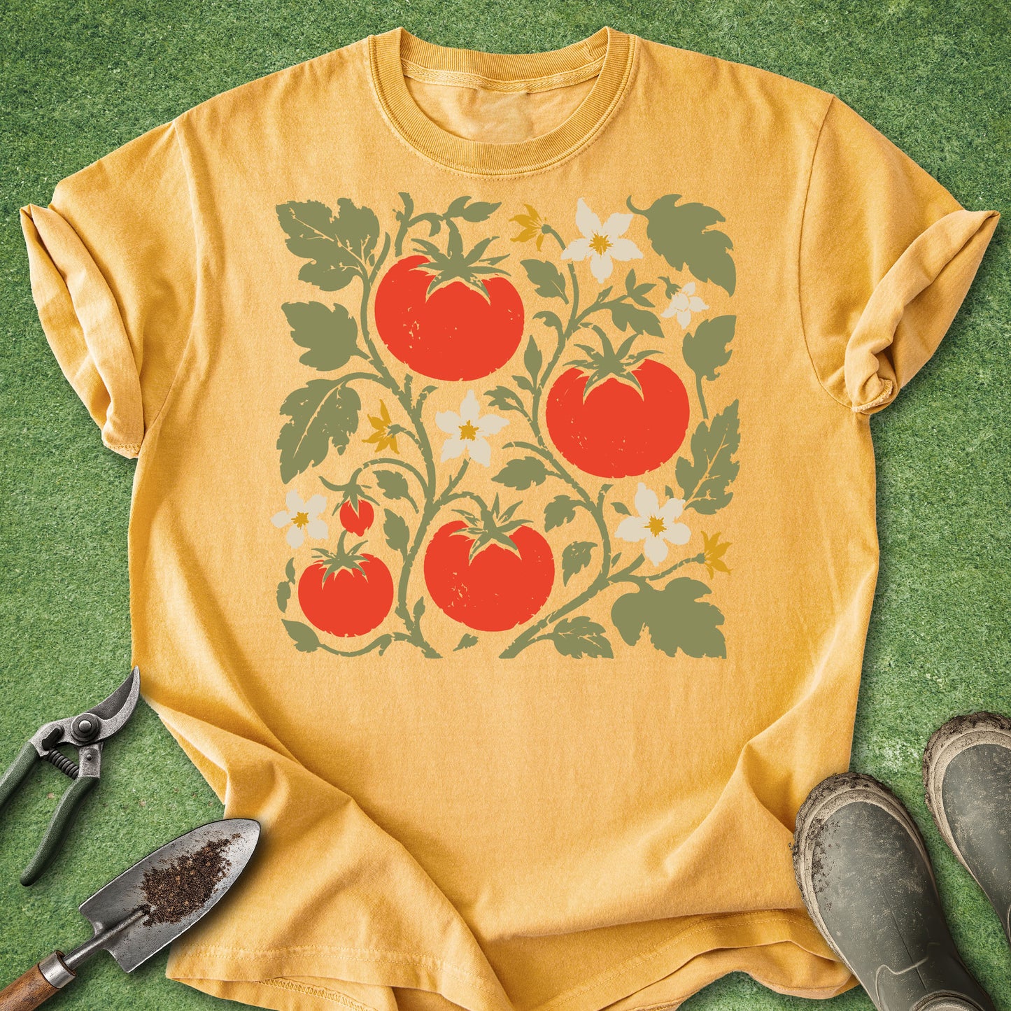 A mustard yellow short-sleeve t-shirt with a retro-style graphic of red tomatoes, green leaves, and white flowers, laid flat on a green grass-like surface with gardening tools and boots nearby.