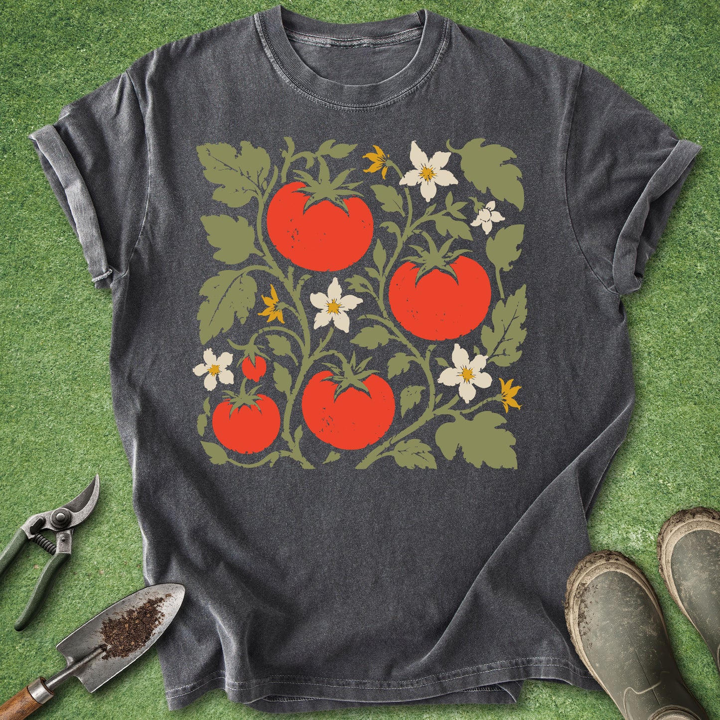 A dark charcoal gray t-shirt laid flat on green grass, featuring a graphic print of red tomatoes, green leaves, and white flowers. Gardening props, including pruning shears, a trowel with soil, and boots, are visible around the shirt.