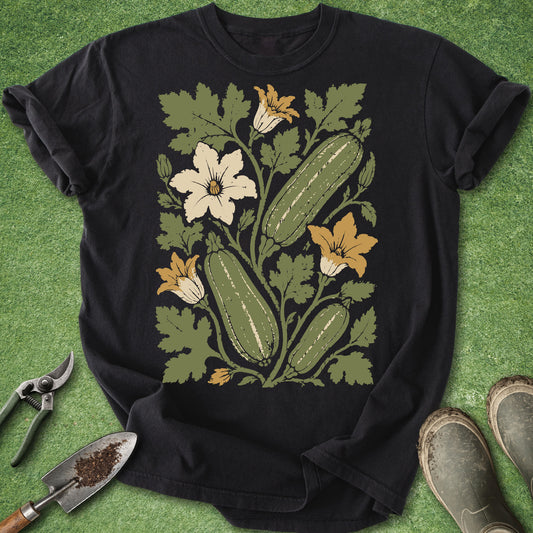 A black short-sleeve graphic T-shirt laid flat on green grass-like ground, featuring a large botanical print of cucumbers, green leaves, and white and yellow flowers. A gardening trowel is at the lower left, and part of a pair of boots is visible at the lower right.