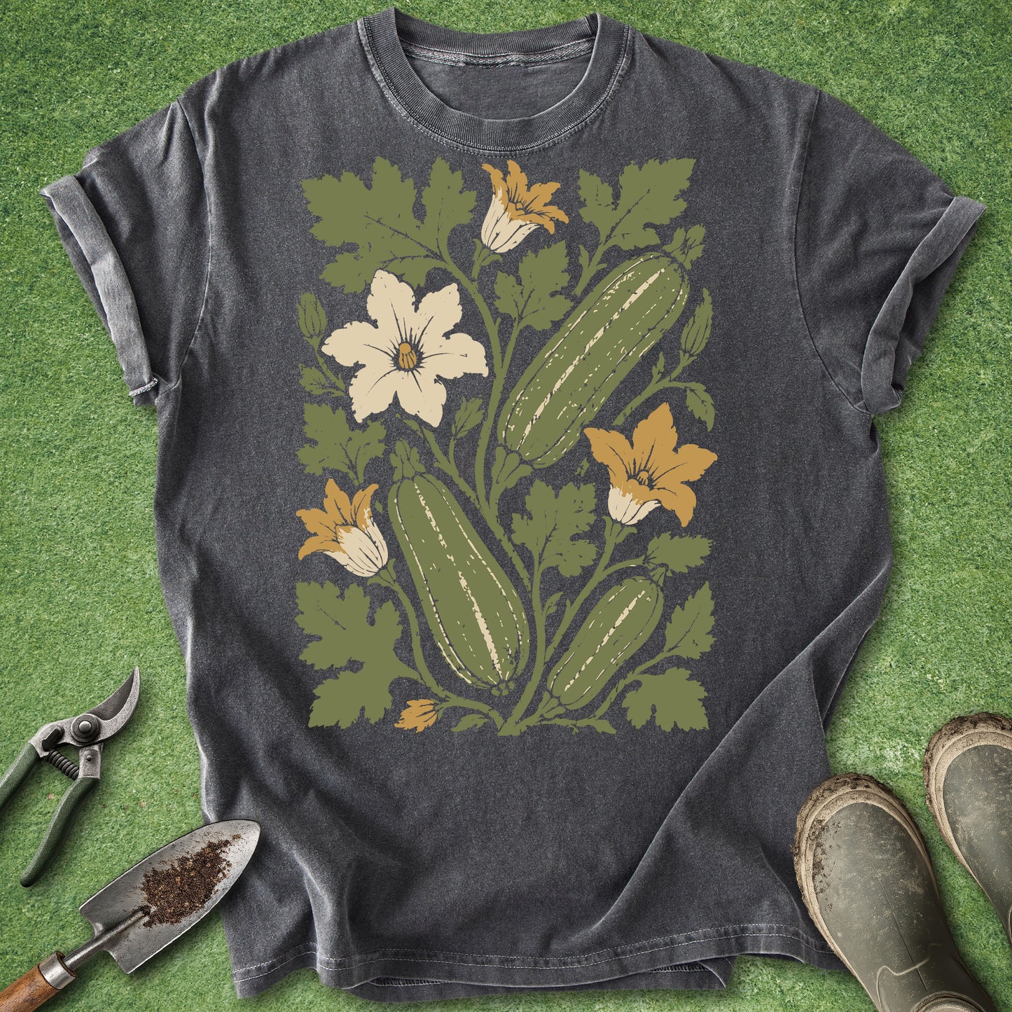 A dark charcoal gray t-shirt with a vintage-style botanical graphic of cucumbers, green leaves, and white and yellow flowers, laid flat on green grass with gardening tools and boots nearby.