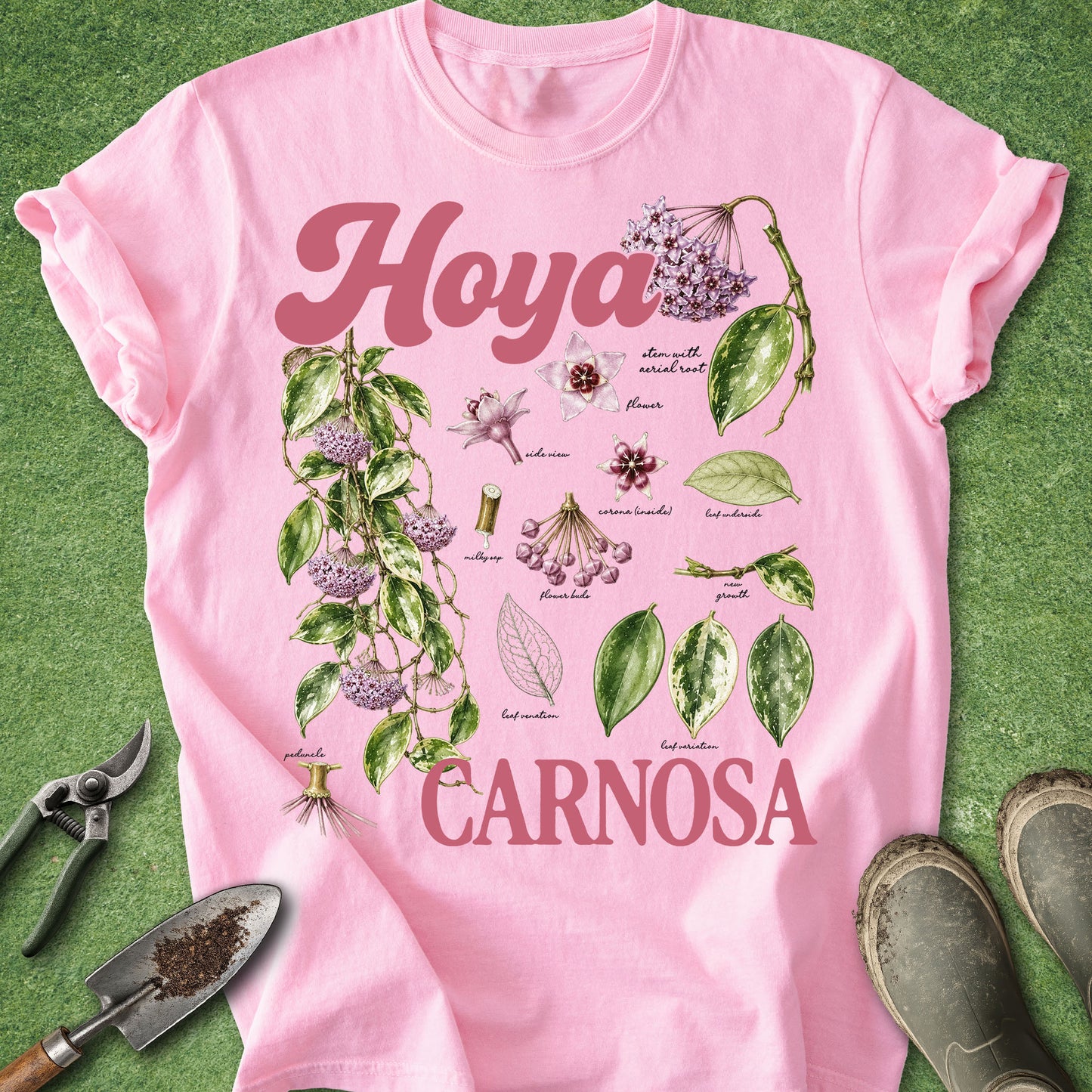 A pink short-sleeve T-shirt laid flat on a green textured surface, featuring botanical illustrations of Hoya carnosa leaves and flowers with the words 'Hoya' and 'Carnosa' in pink lettering. Gardening tools and shoes are placed around the shirt as styling props.