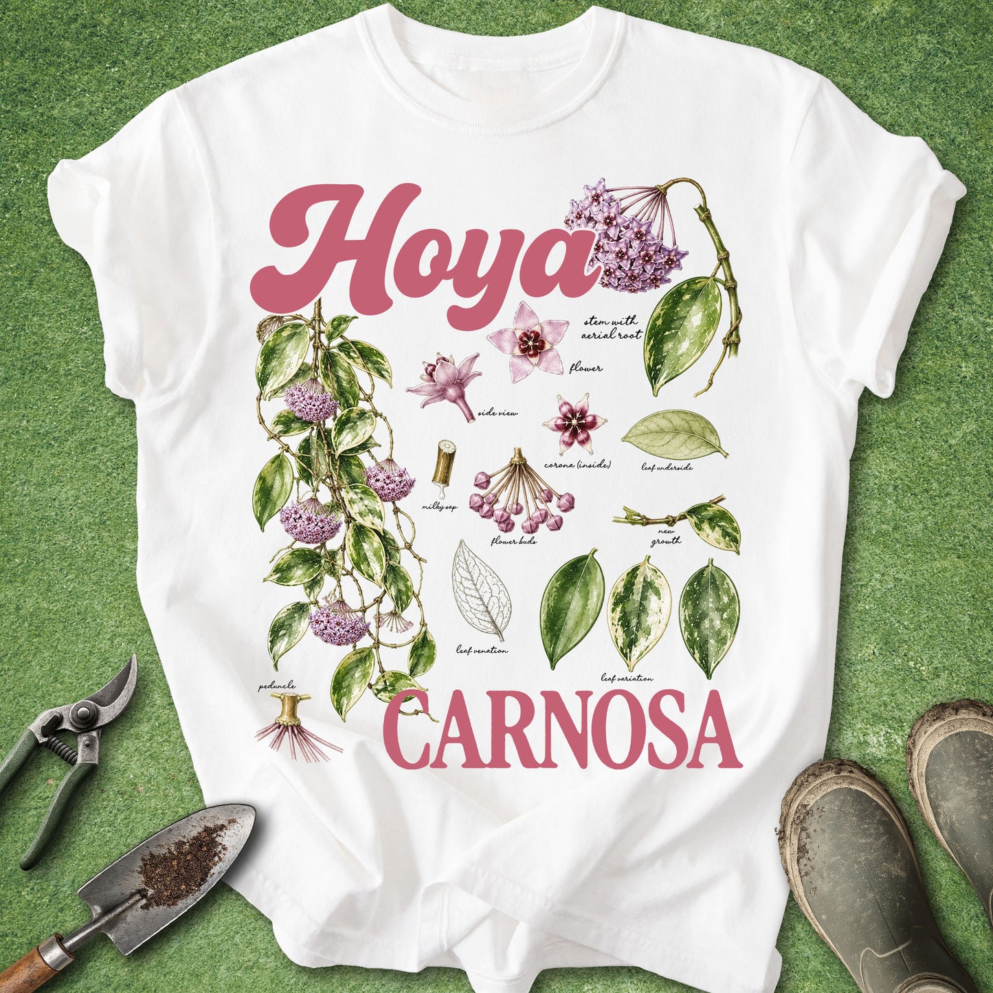 A white short-sleeve T-shirt laid flat on green grass, featuring a botanical-style print with the words 'Hoya Carnosa' in pink, illustrated purple flowers, green leaves, and small botanical labels. Gardening tools and boots are placed around the shirt as styling props.