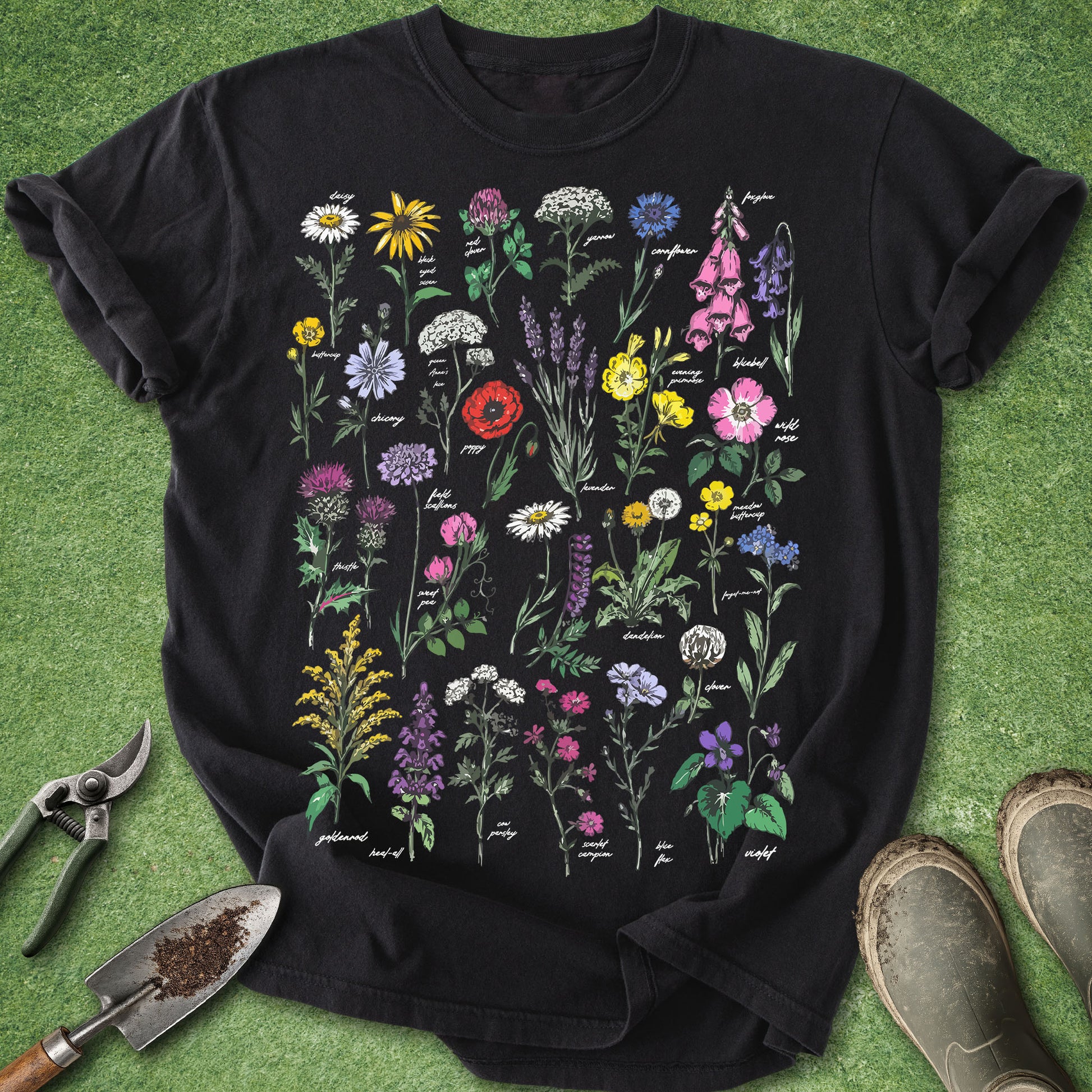 Black Comfort Colors t-shirt featuring a colorful collection of wildflowers in a vintage botanical field guide style with labeled blooms including lavender, poppy, foxglove, clover, daisies, and violets.