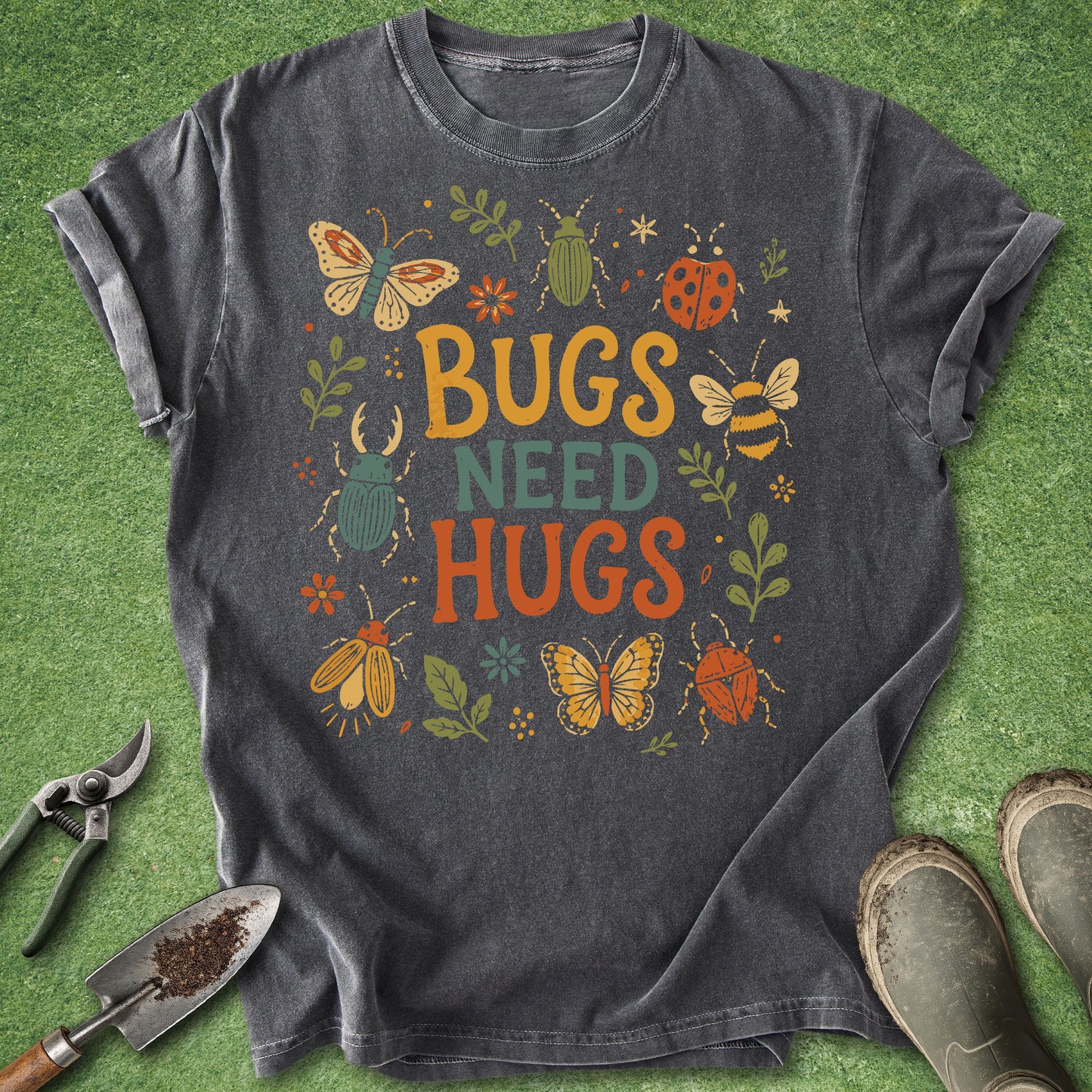 A dark gray graphic t-shirt laid flat on a green grass-like surface, surrounded by gardening props including pruning shears, a trowel, and boots. The shirt features a colorful retro-style design with the text 'BUGS NEED HUGS' surrounded by illustrated butterflies, bees, ladybugs, and flowers.