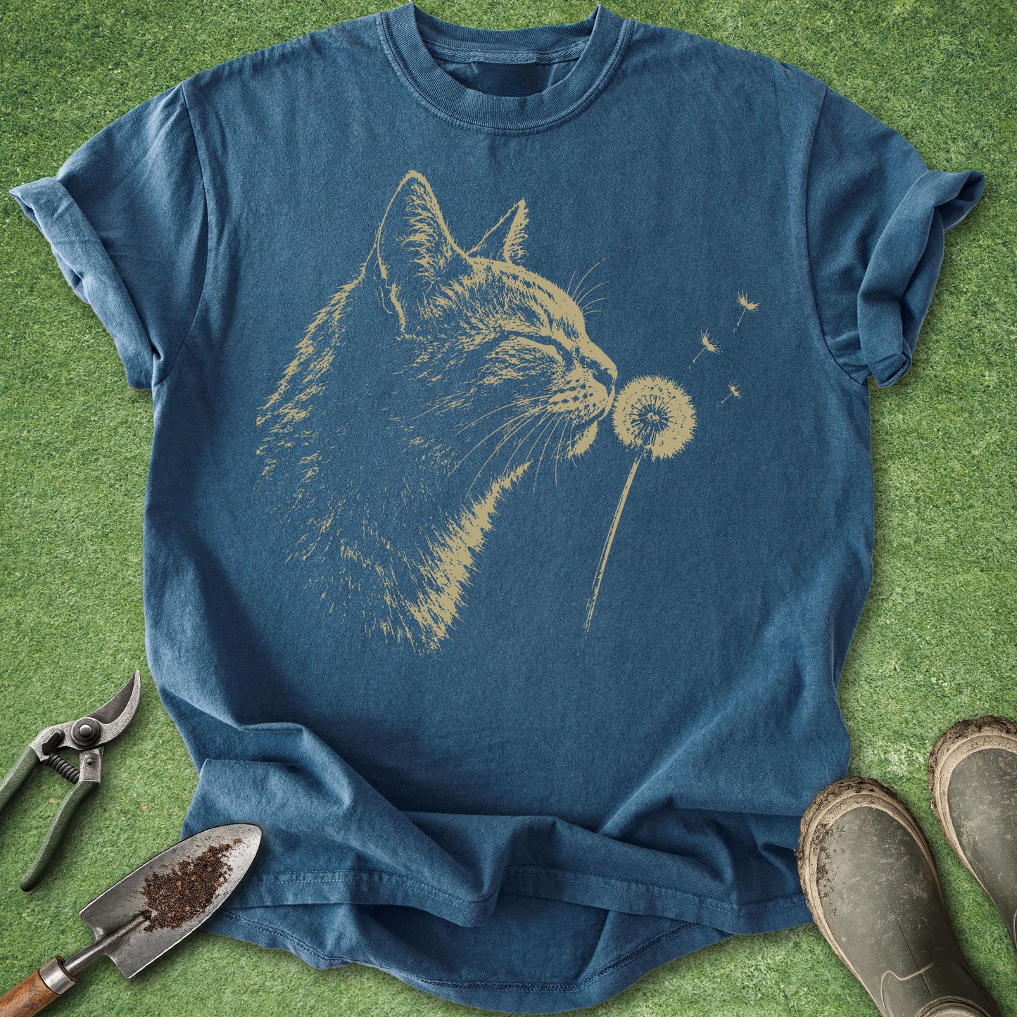 Blue t-shirt with a cat blowing on a dandelion graphic on a grassy background.