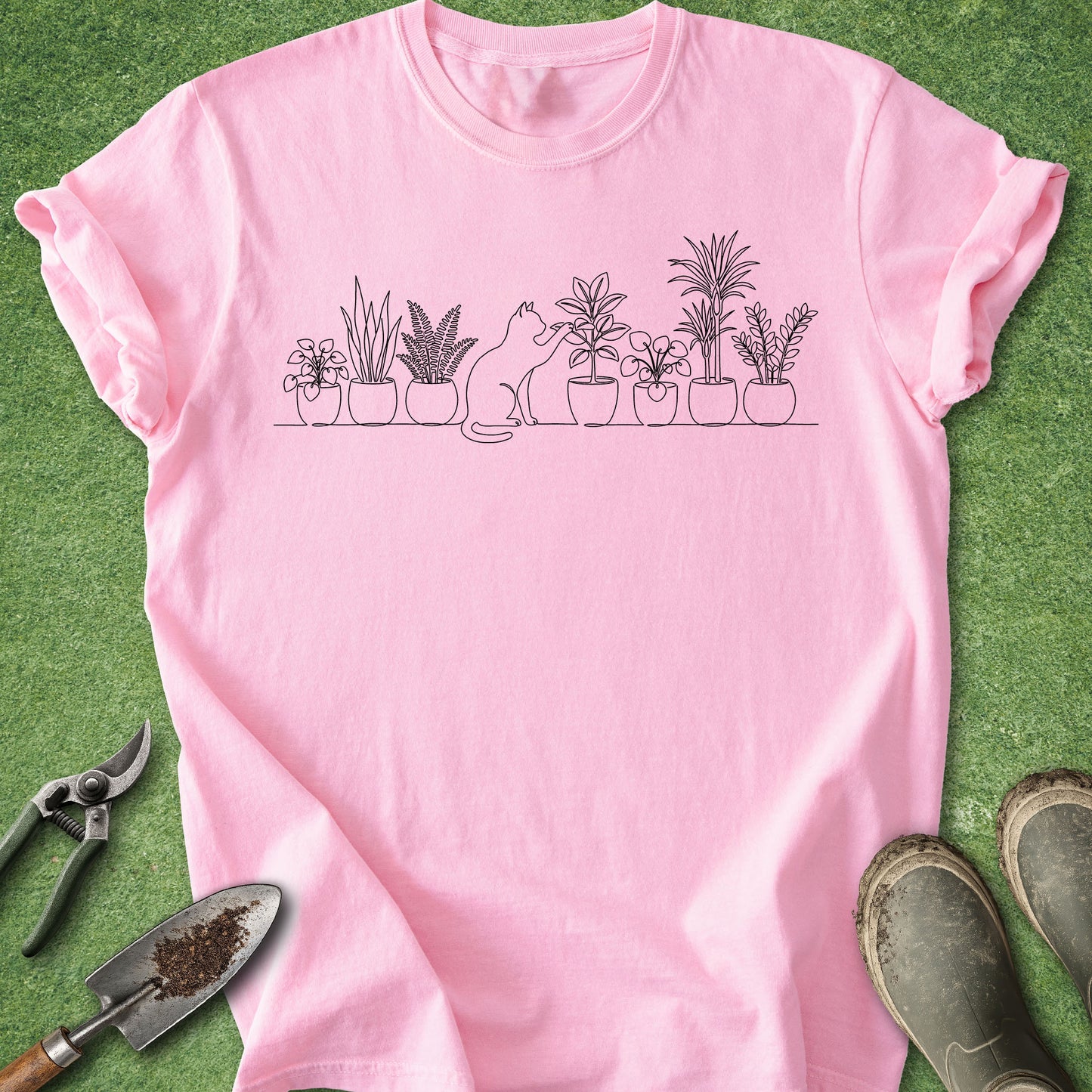 A pink short-sleeve t-shirt with a black line art graphic of potted plants and a cat, laid flat on green grass with gardening tools and boots nearby.