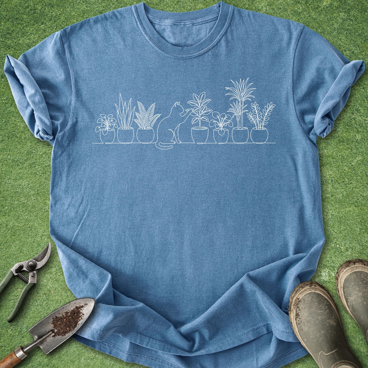 A blue crewneck t-shirt with a white line art graphic of potted plants and a cat, laid flat on green grass with gardening tools and boots nearby.