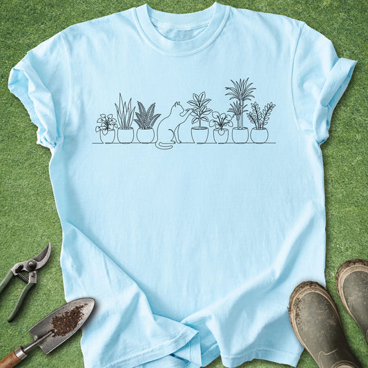 A light blue short-sleeve t-shirt laid flat on green grass, featuring a black line art graphic of potted plants and a cat. Gardening tools and brown boots are placed around the shirt as props.