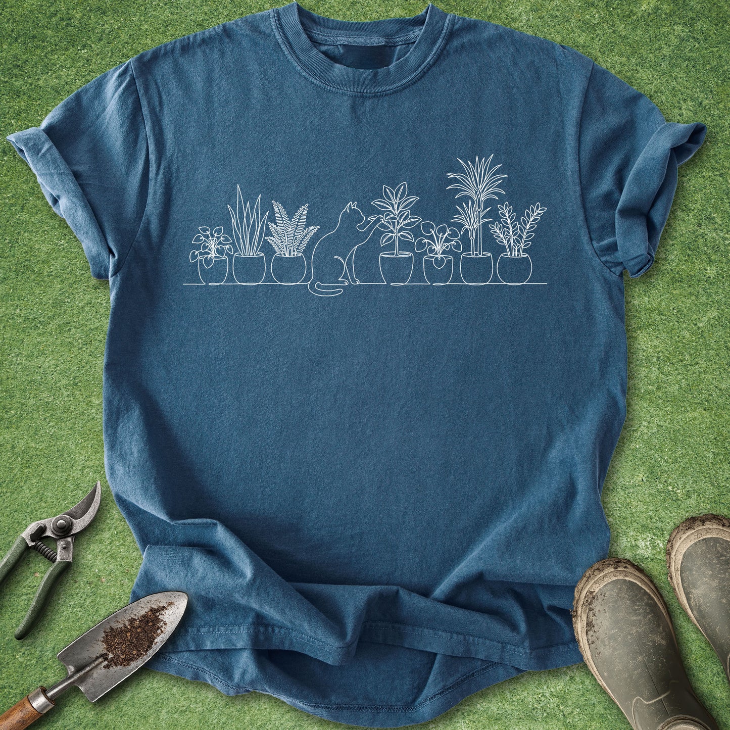 A blue crewneck t-shirt with a white line art graphic of a cat and potted plants, laid flat on green grass with gardening tools and boots nearby.