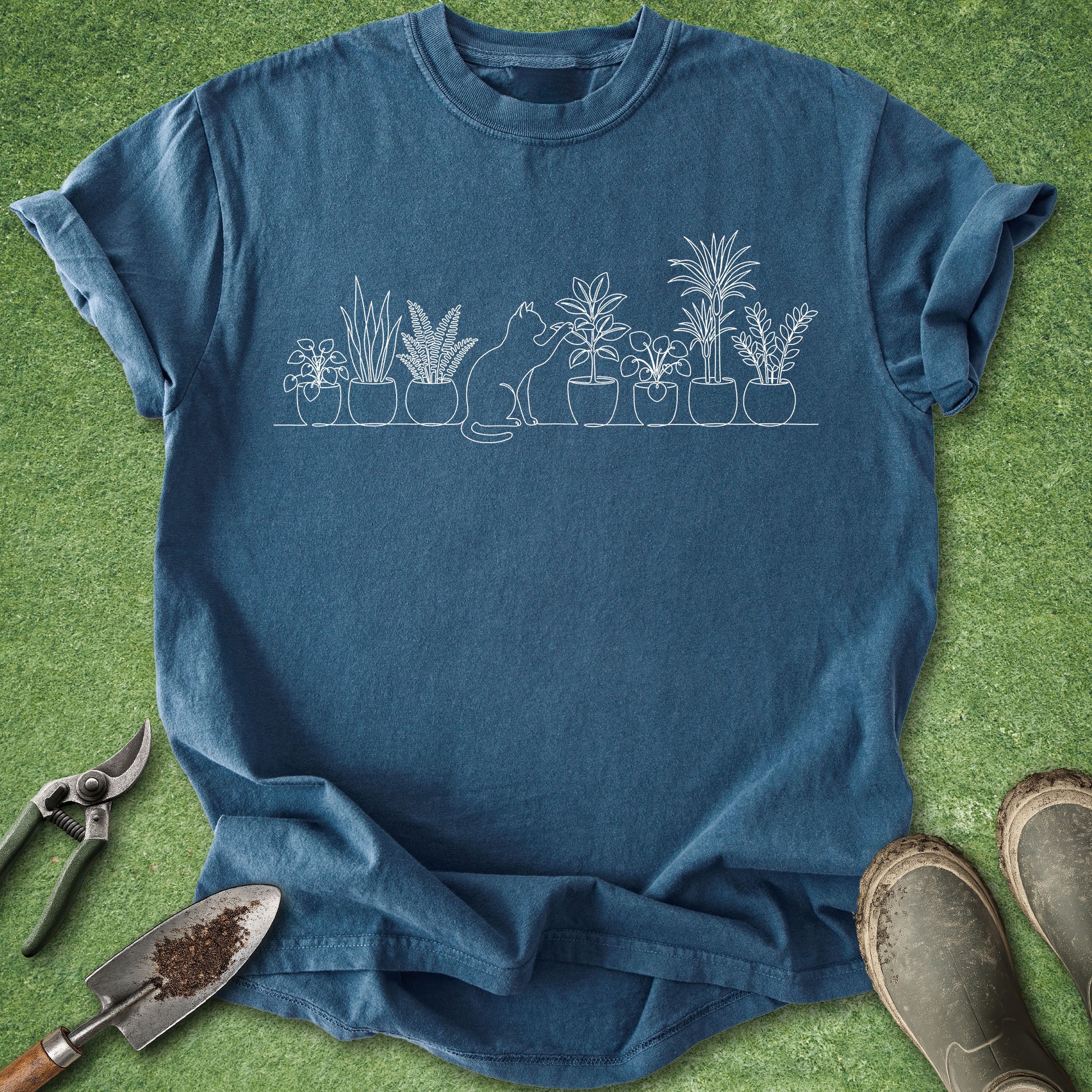 A blue crewneck t-shirt with a white line art graphic of a cat and potted plants, laid flat on green grass with gardening tools and boots nearby.