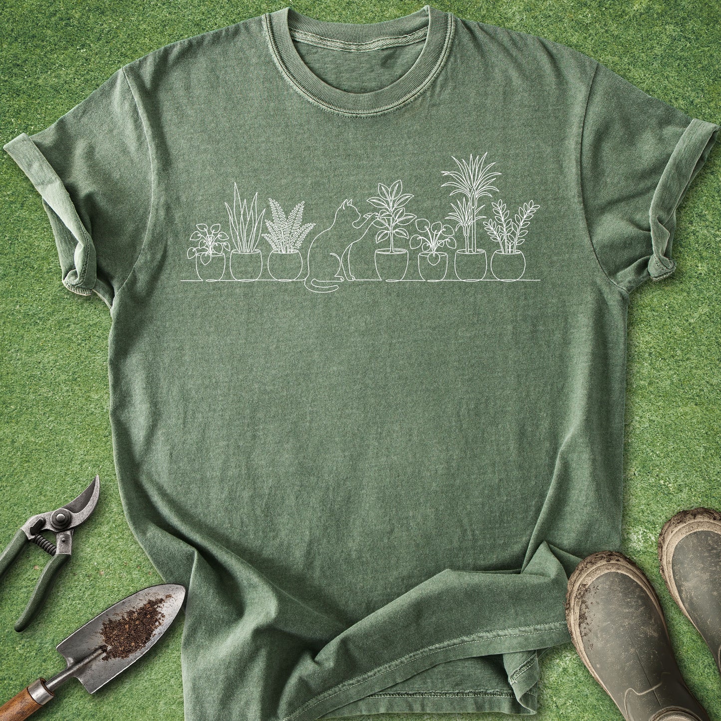 An olive green heathered t-shirt with a white line art graphic of a cat and potted plants, laid flat on green grass with gardening props (trowel, pruning shears, boots) around it.