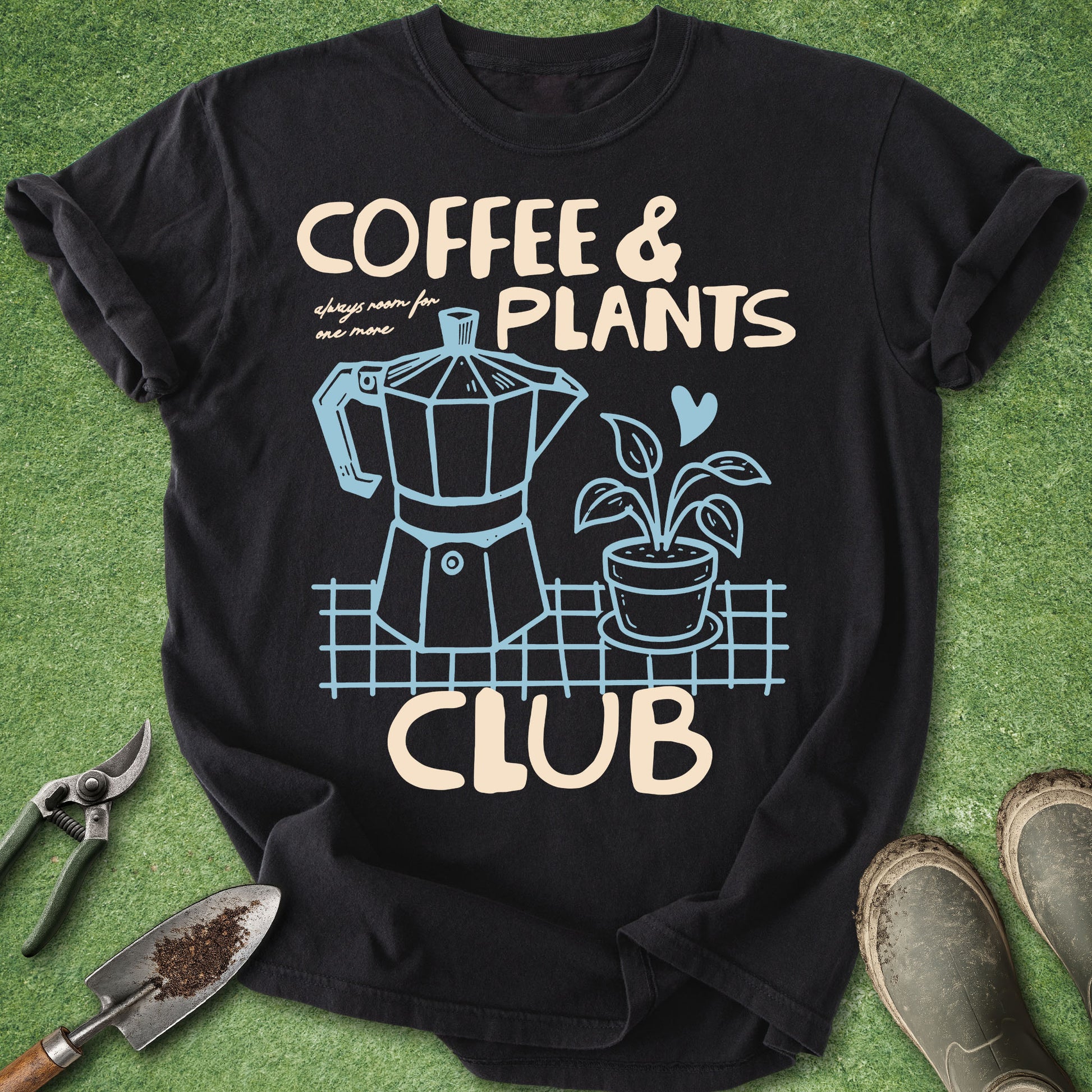 A black graphic t-shirt laid flat on green grass-like artificial turf, with gardening props around it. The shirt has a cream and light blue print reading 'COFFEE & PLANTS CLUB' with an illustrated moka pot and potted plant.