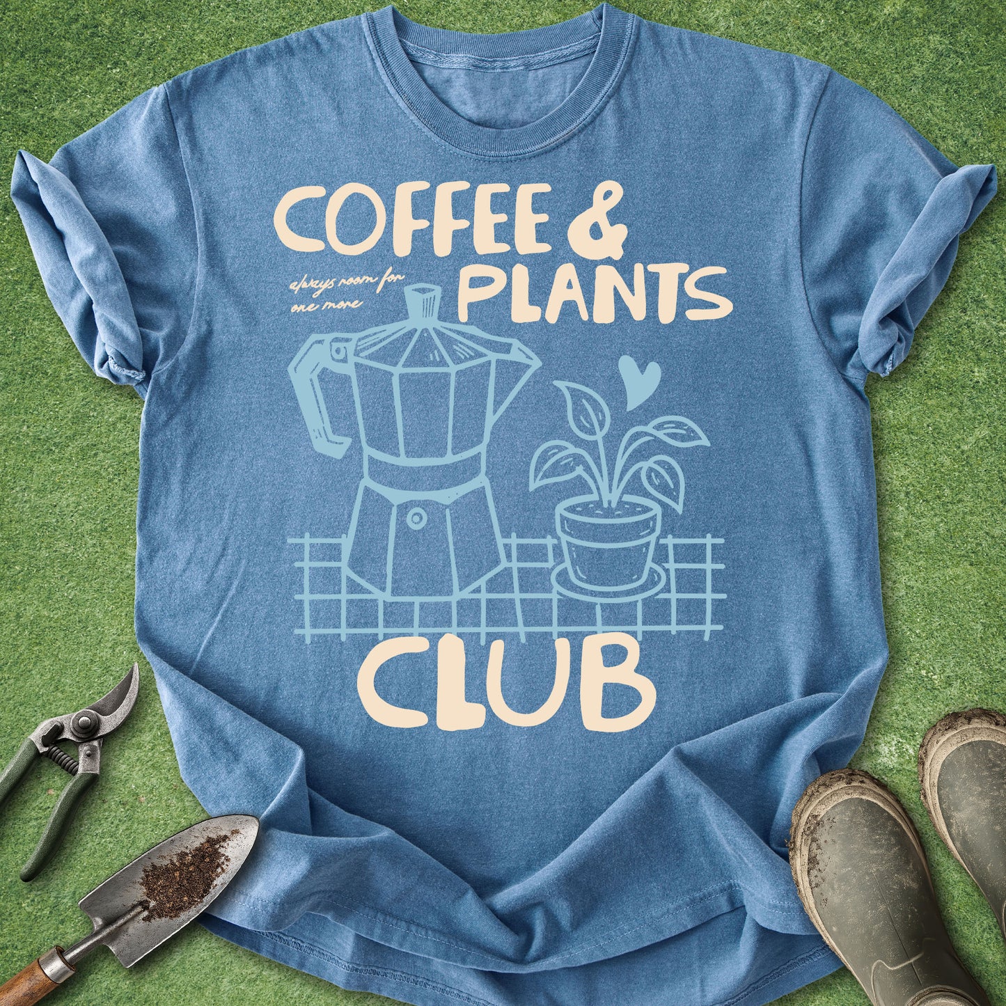 A blue graphic t-shirt laid flat on green grass with gardening props, featuring the text 'COFFEE & PLANTS CLUB' and illustrations of a moka pot and a potted plant.