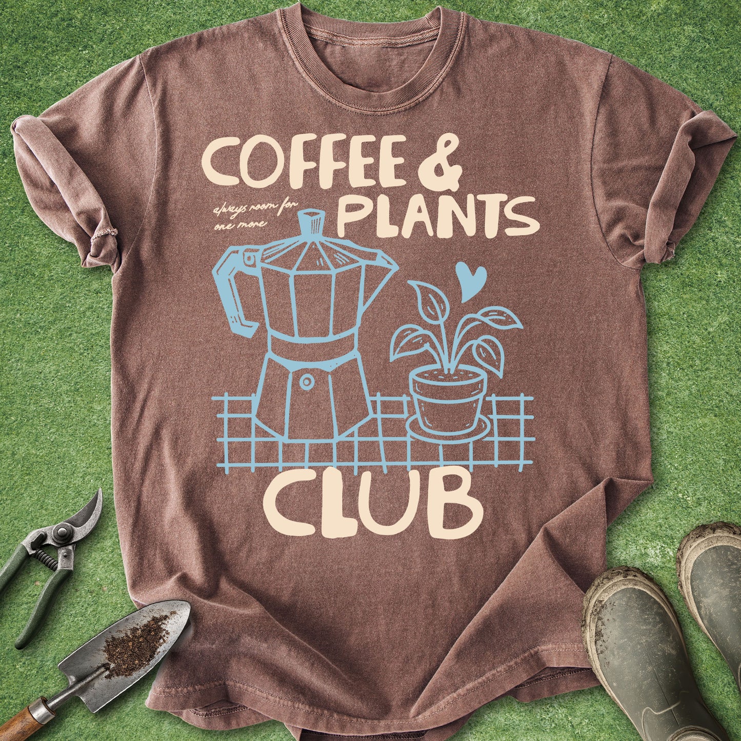 A brown short-sleeve graphic T-shirt laid flat on green grass-like background, with gardening tools and muddy boots around it. The shirt reads "COFFEE & PLANTS CLUB" with a coffee maker and potted plant illustration in light beige and blue.