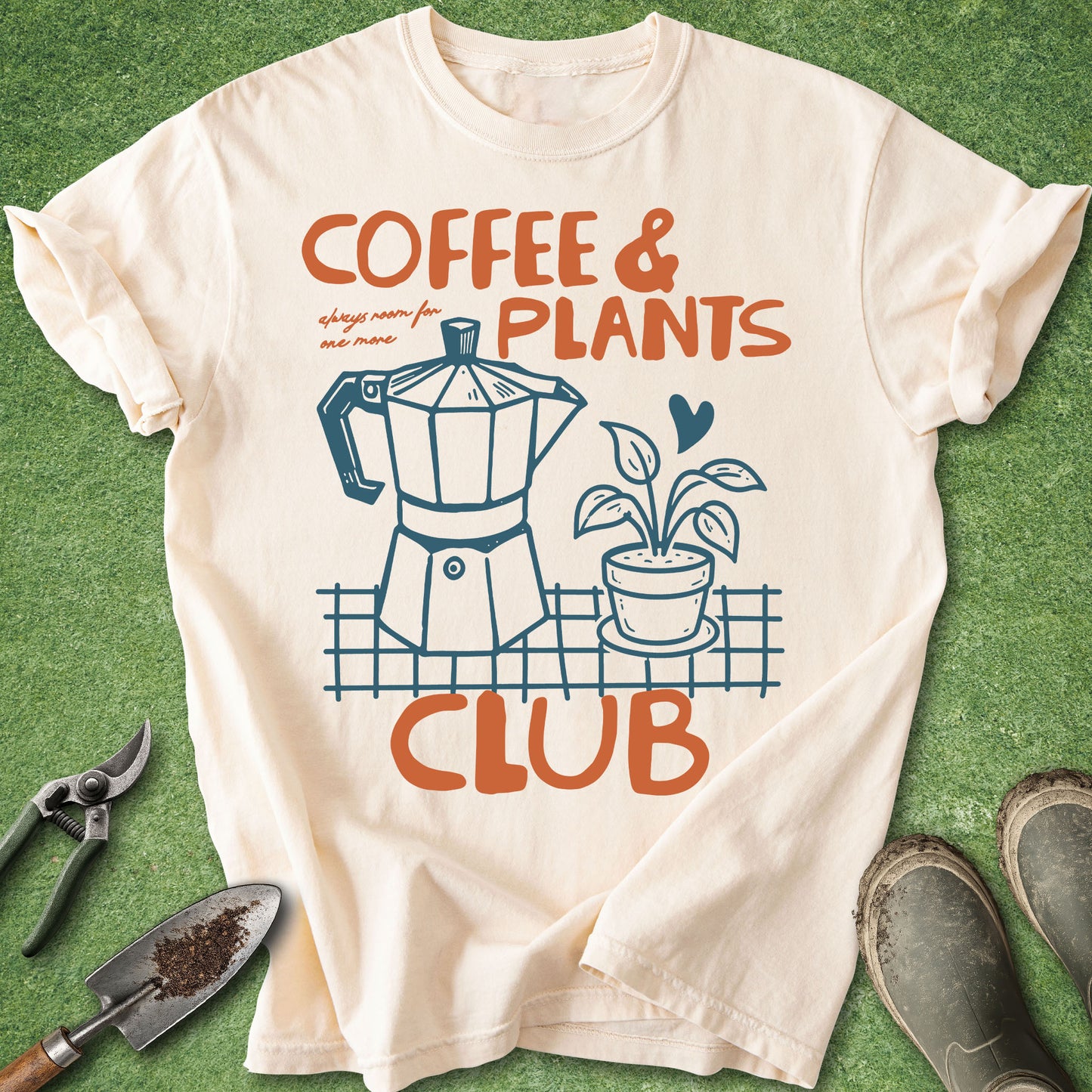 An off-white graphic t-shirt laid flat on green grass with gardening props, featuring the text 'COFFEE & PLANTS CLUB' and an illustration of a coffee maker and a potted plant.