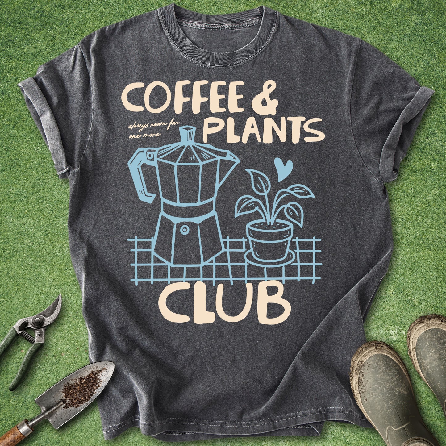 A dark charcoal gray graphic t-shirt laid flat on green grass-like artificial turf, surrounded by gardening props including pruning shears, a trowel, and muddy boots. The shirt features a cream-colored and light blue graphic of a moka pot and a potted plant with the text 'COFFEE & PLANTS CLUB'.