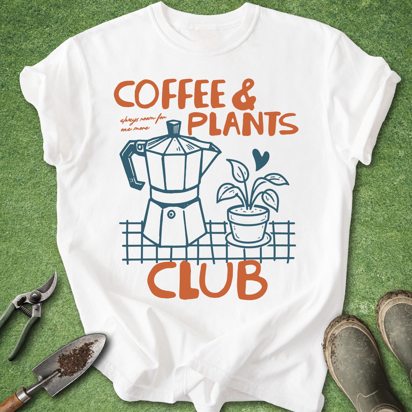 A white graphic t-shirt laid flat on green grass with gardening props around it. The shirt features an orange and blue graphic that reads 'COFFEE & PLANTS CLUB' with an illustration of a moka pot and a potted plant.