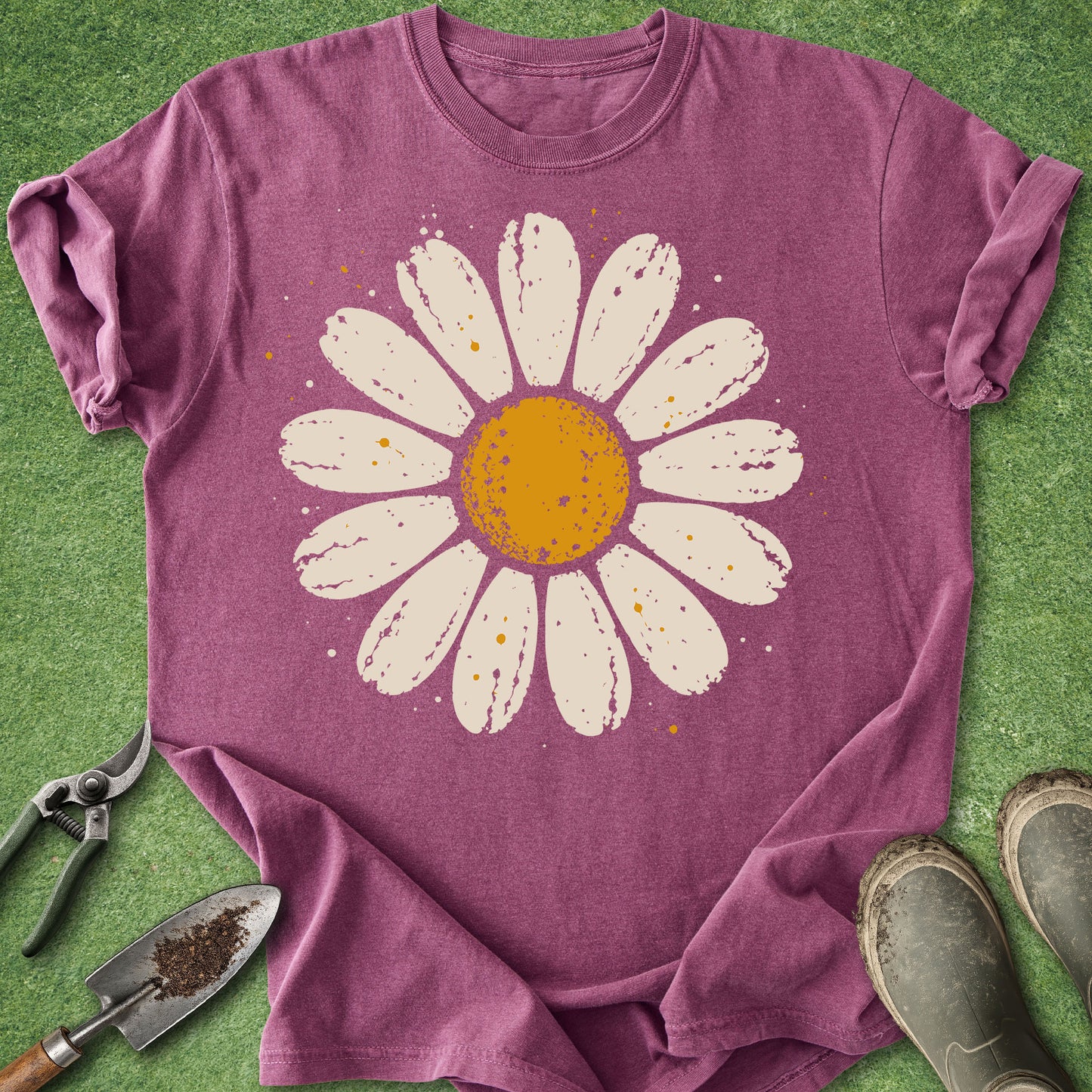 A heathered mauve purple t-shirt laid flat on green grass, featuring a large distressed white daisy graphic with a yellow-orange center. Gardening props including a trowel, pruning shears, and boots are visible around the shirt.