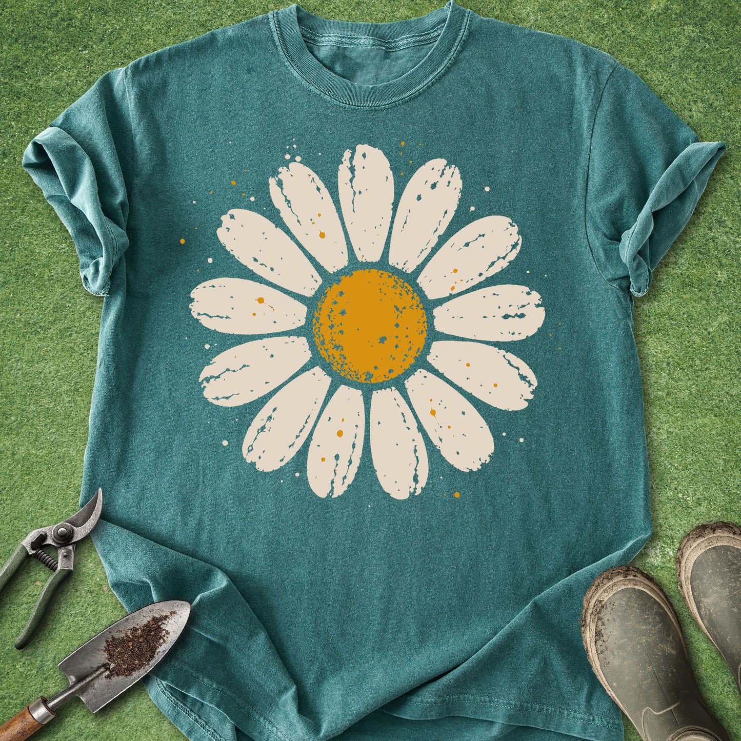 A teal short-sleeve t-shirt with a large white daisy graphic featuring a yellow-orange center, laid flat on green grass with gardening tools and boots nearby.
