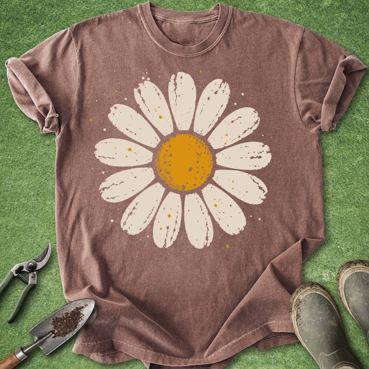 A brown short-sleeve t-shirt with a large distressed white daisy graphic featuring a yellow-orange center, laid flat on green artificial turf with gardening props like pruning shears, a trowel, and boots.