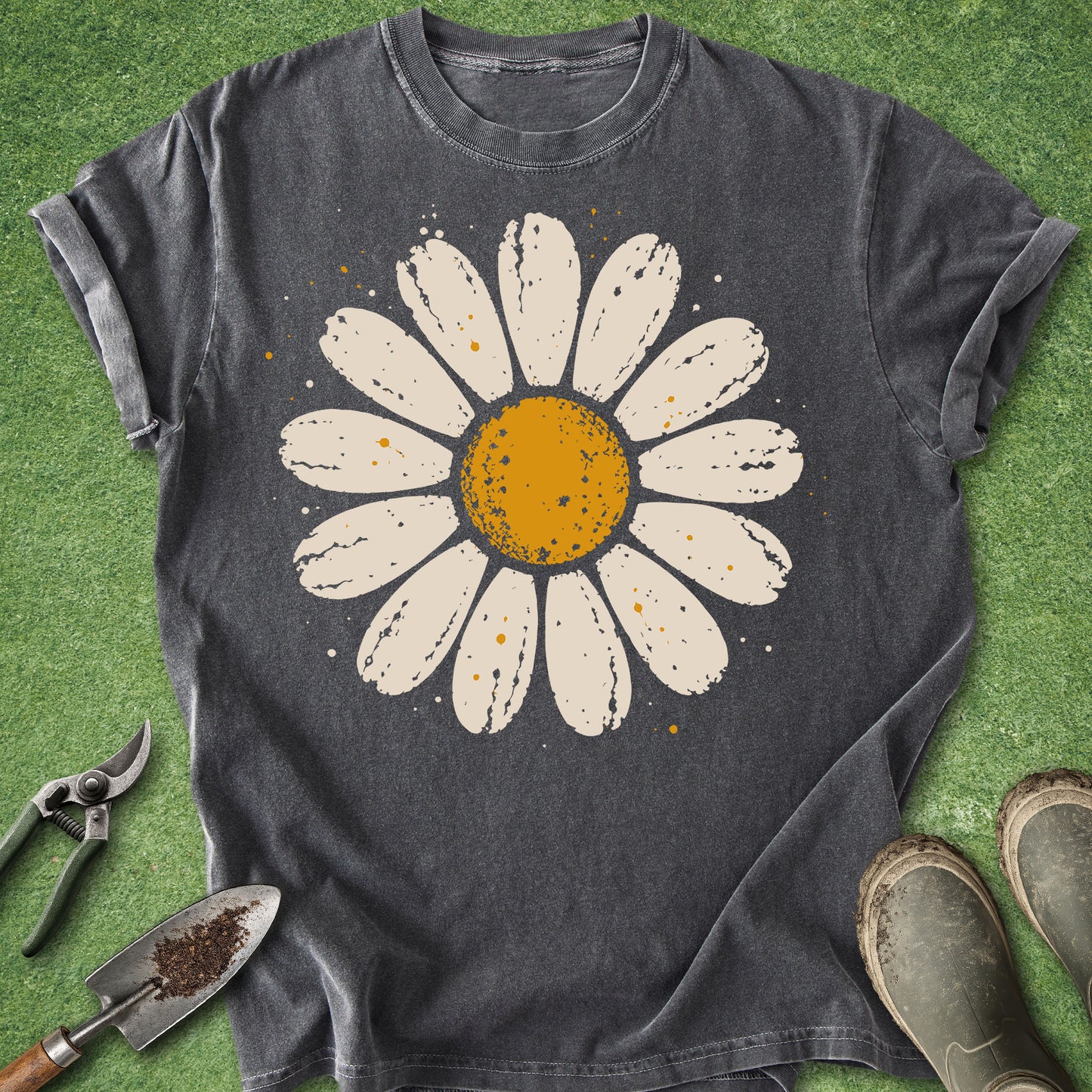 A dark charcoal gray t-shirt with a distressed white daisy graphic featuring a yellow-orange center, laid flat on green artificial turf with gardening tools and boots nearby.