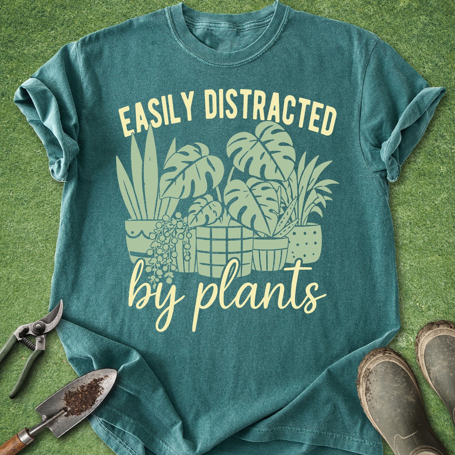 Teal t-shirt with plant-themed design and gardening tools on a grass background