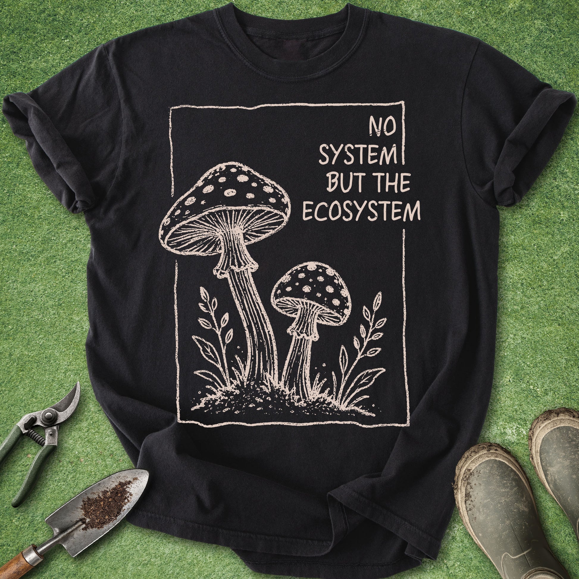Black t-shirt with mushroom design and 'No System But The Ecosystem' text on a green surface with gardening tools.