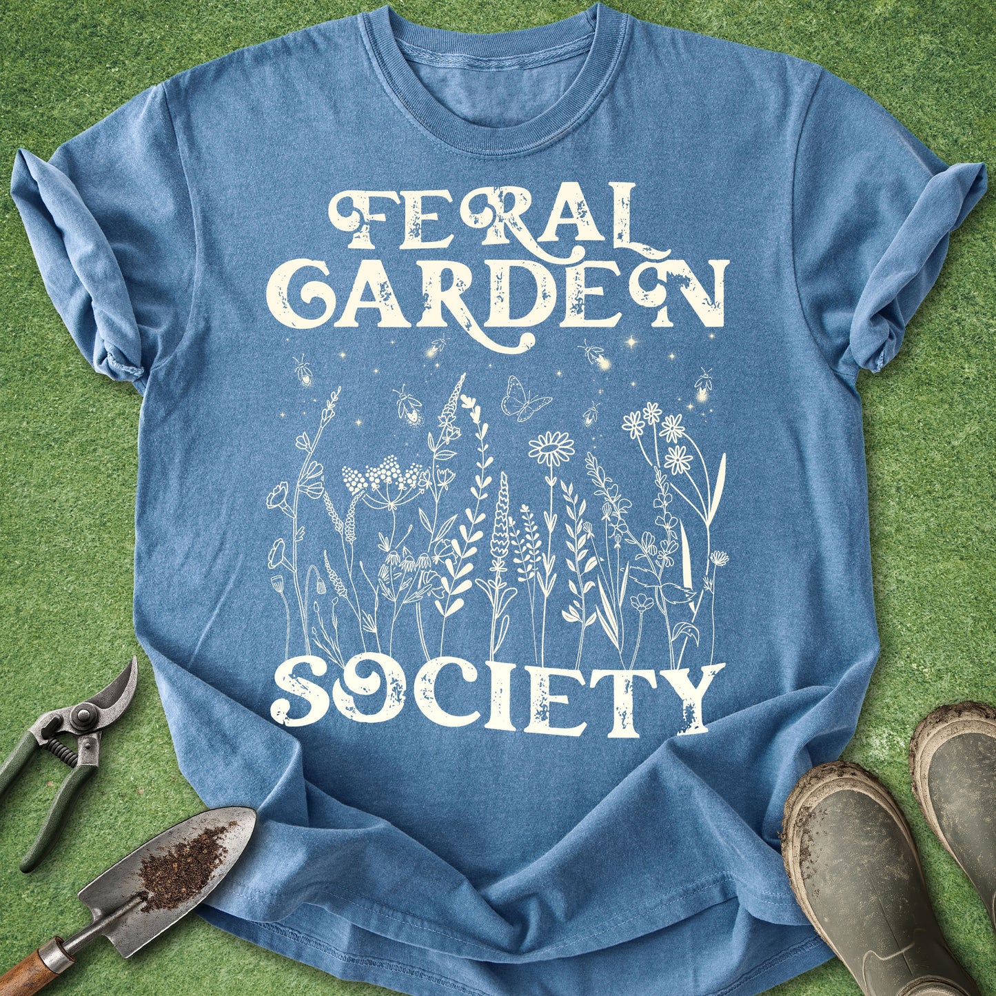 A blue short-sleeve graphic T-shirt laid flat on green grass-like ground, with white text reading 'FERAL GARDEN SOCIETY' and white botanical line art across the front. Gardening props including pruning shears, a trowel, and boots are partially visible around the shirt.