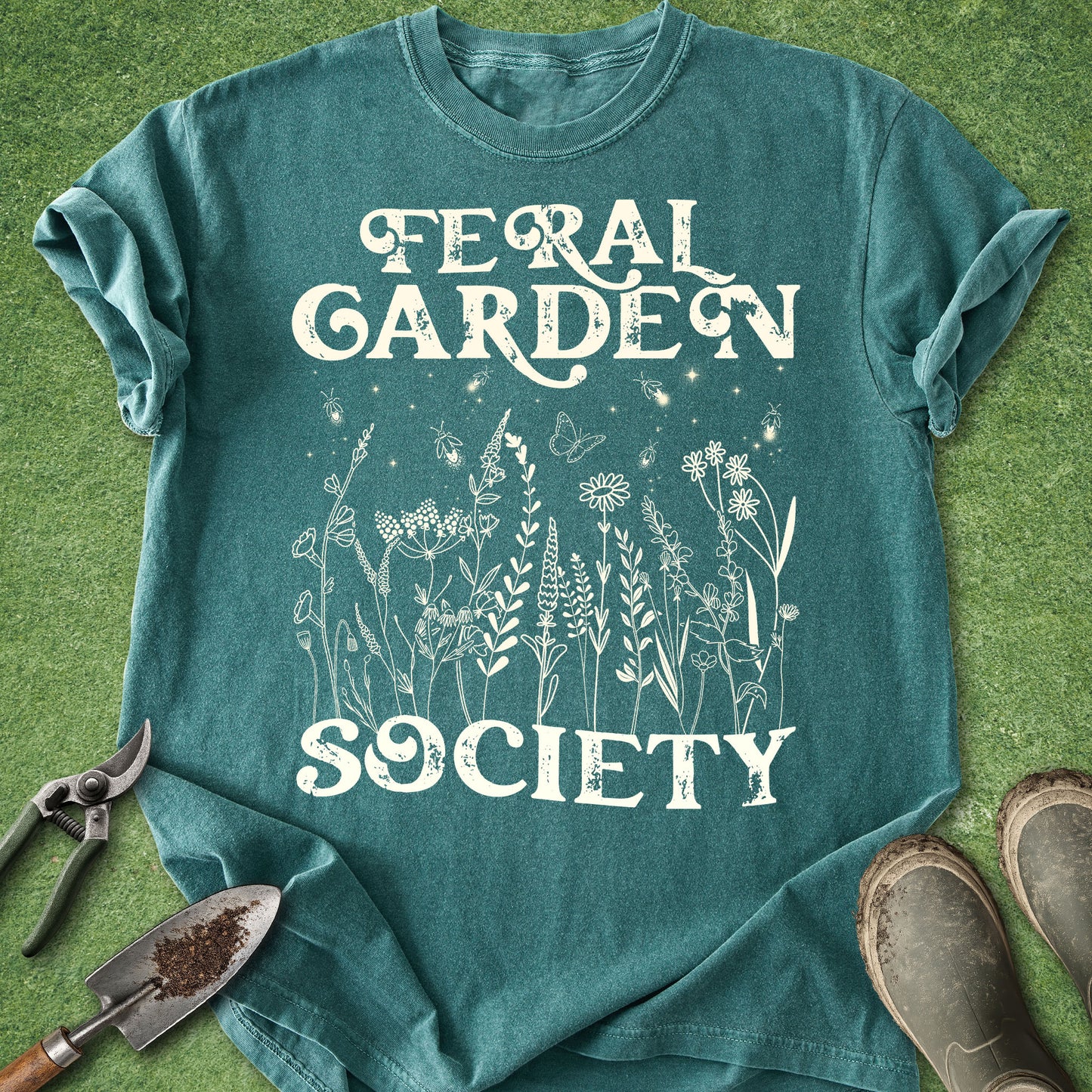 A teal t-shirt with a white graphic reading 'Feral Garden Society' and featuring wildflower illustrations, laid flat on green grass with gardening tools and boots nearby.