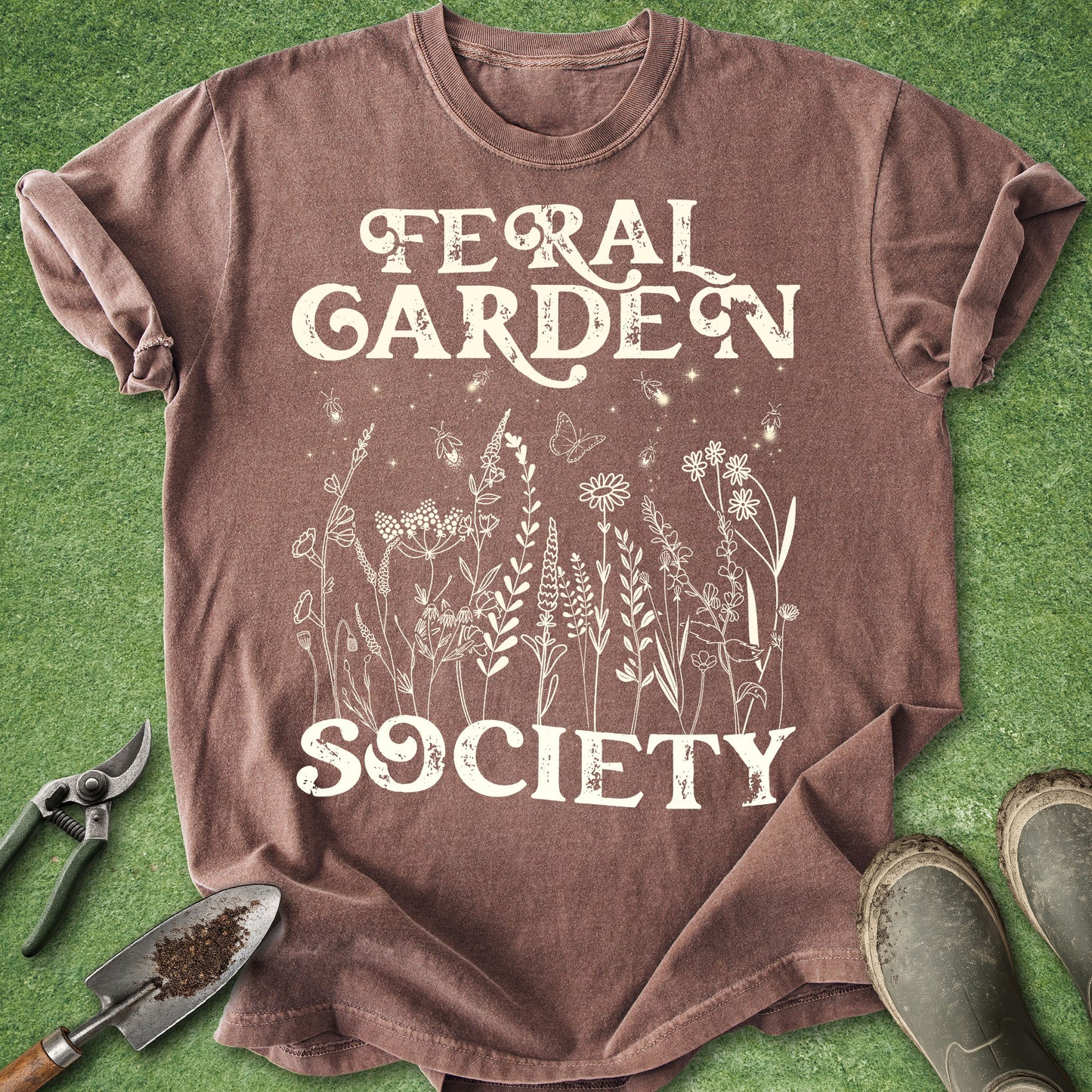 A brown short-sleeve graphic T-shirt laid flat on green grass-like ground, with white text reading 'Feral Garden Society' and a white botanical illustration on the front. Gardening props including pruning shears, a trowel, and boots are placed around the shirt.