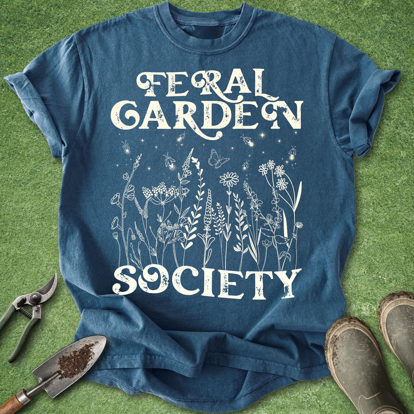 A blue graphic t-shirt laid flat on green grass with gardening props, featuring the white text 'FERAL GARDEN SOCIETY' and a white line-art floral design.