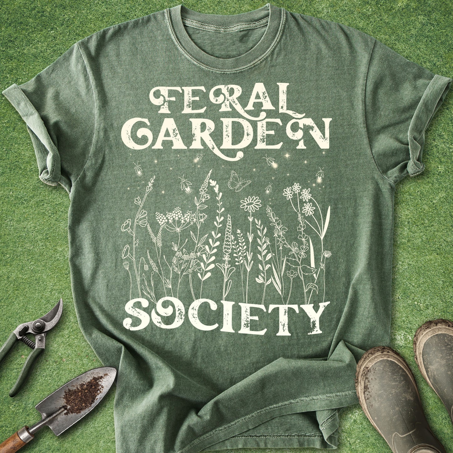 An olive green heathered graphic t-shirt laid flat on artificial green grass, featuring the words 'FERAL GARDEN SOCIETY' in a distressed white font with white line drawings of garden plants and butterflies. Gloves, a trowel, and pruning shears are styled around the shirt.
