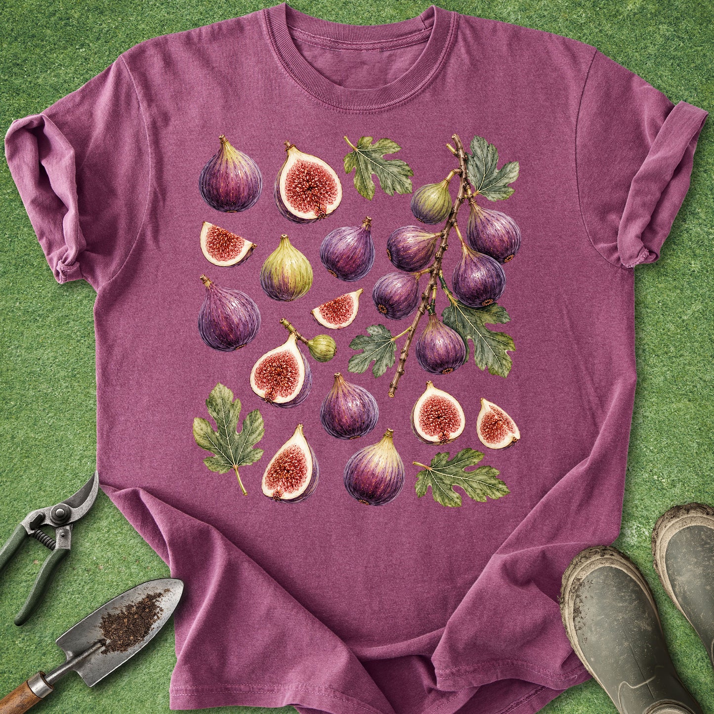 A mauve-purple short-sleeve t-shirt with a detailed graphic print of various figs (whole and sliced) with green leaves, laid flat on a green grass-like surface. Gardening tools (trowel, pruning shears) and muddy boots are visible as props around the shirt.