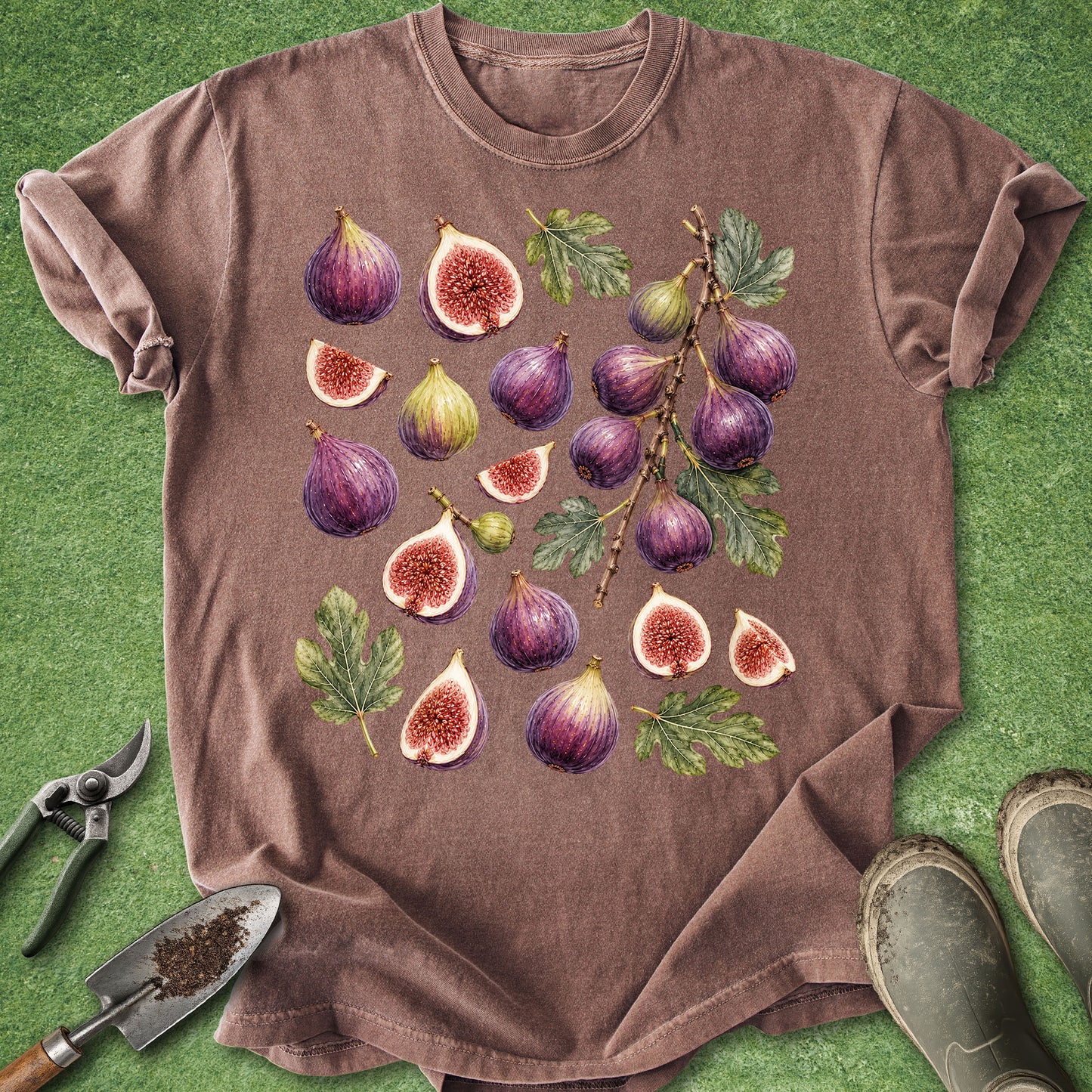 A brown short-sleeve t-shirt laid flat on green grass, featuring a large graphic print of illustrated purple and green figs with leaves. Gardening tools and muddy boots are placed around the shirt as styling props.