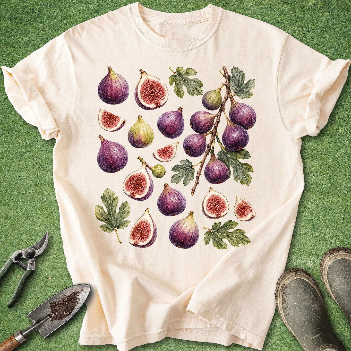 An off-white short-sleeve t-shirt with a detailed botanical print of purple and green figs, laid flat on green artificial turf with gardening props like pruning shears, a trowel, and boots.