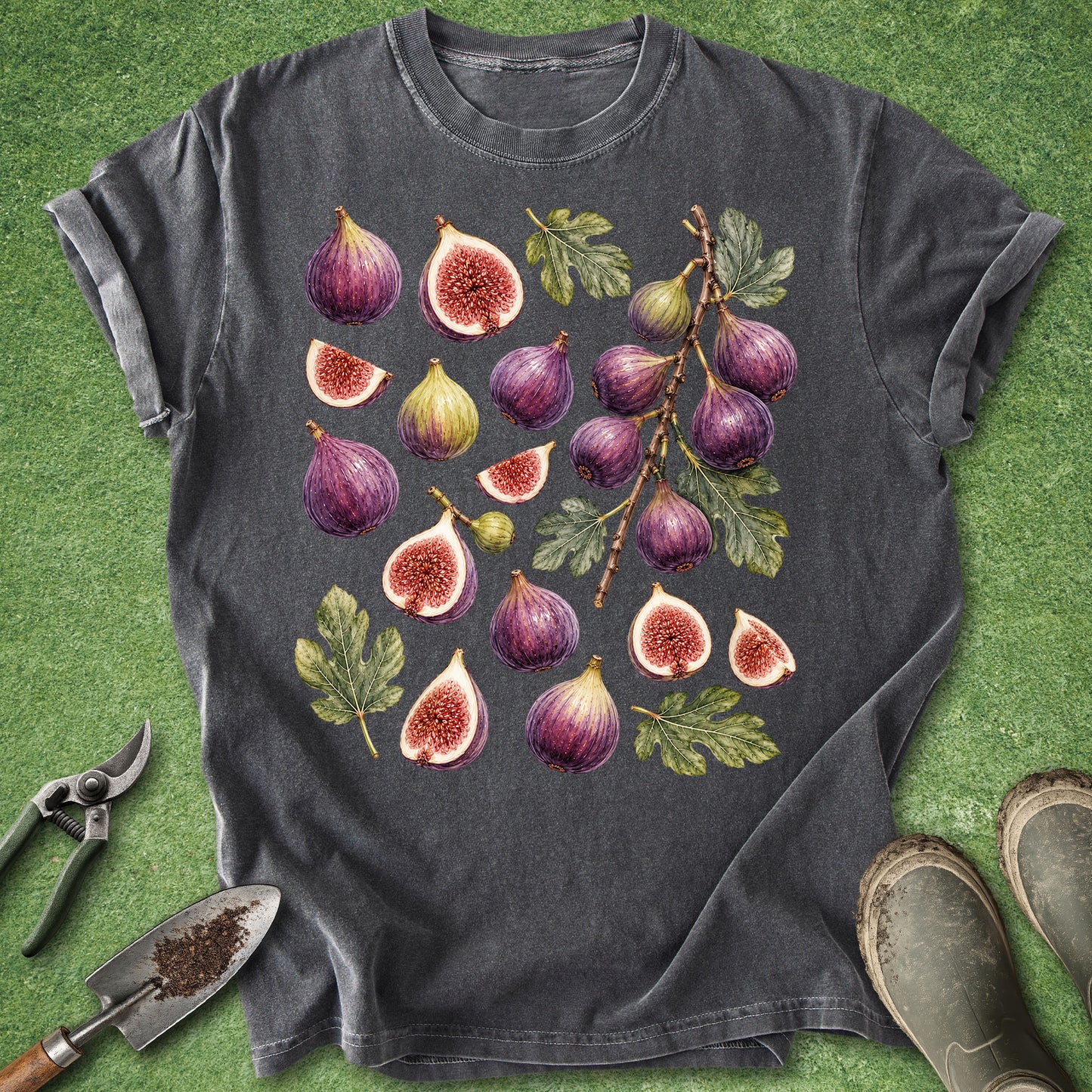 A dark gray t-shirt with a detailed watercolor print of various figs and leaves, styled flat on a green grass-like background with gardening tools and boots.
