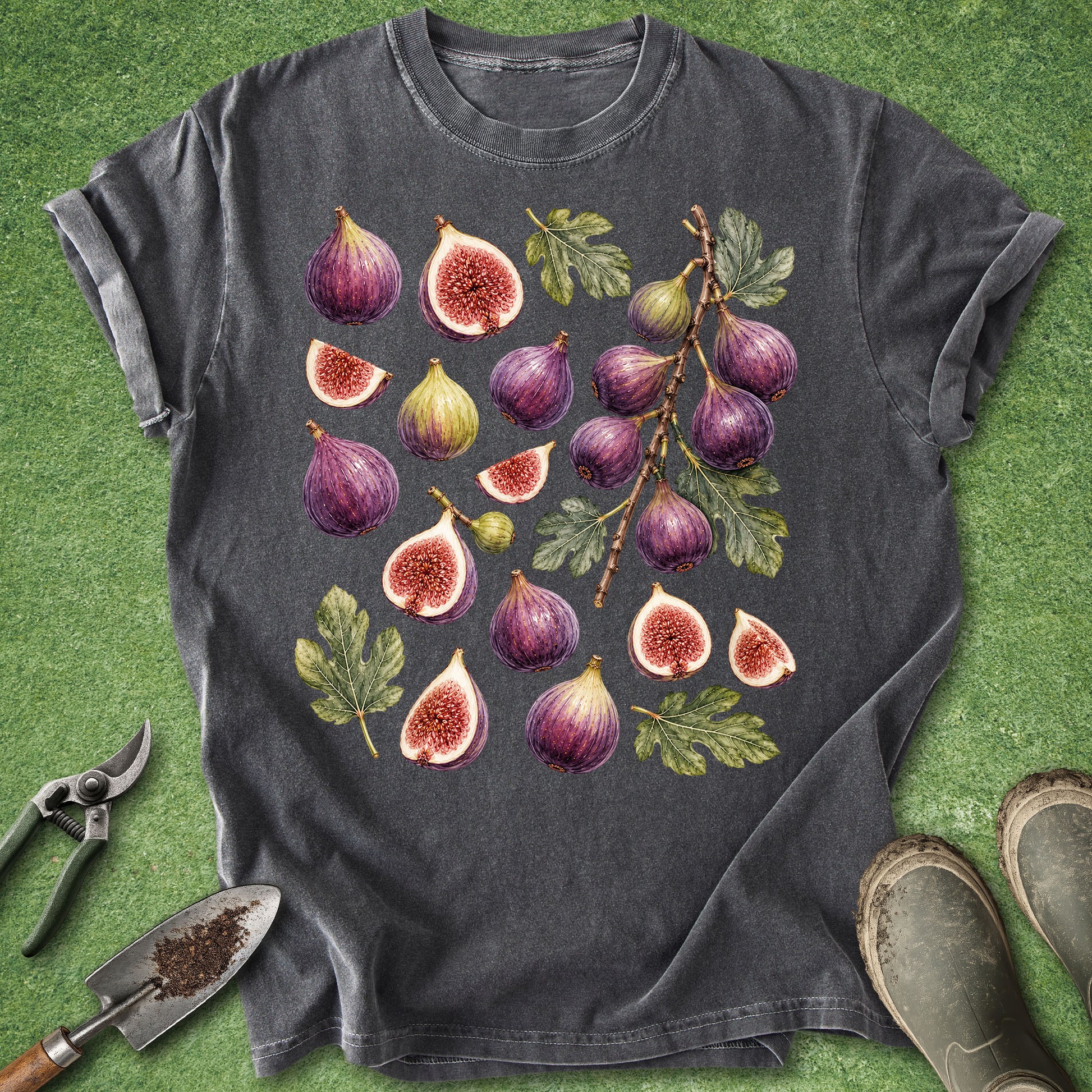 A dark gray t-shirt with a detailed watercolor print of various figs and leaves, styled flat on a green grass-like background with gardening tools and boots.