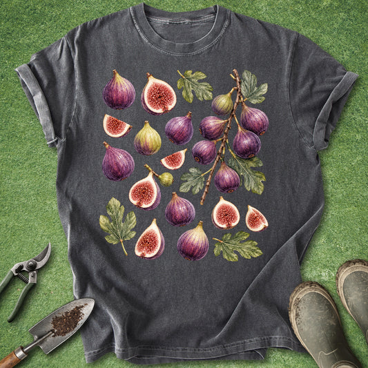 A dark gray t-shirt with a detailed watercolor print of various figs and leaves, styled flat on a green grass-like background with gardening tools and boots.