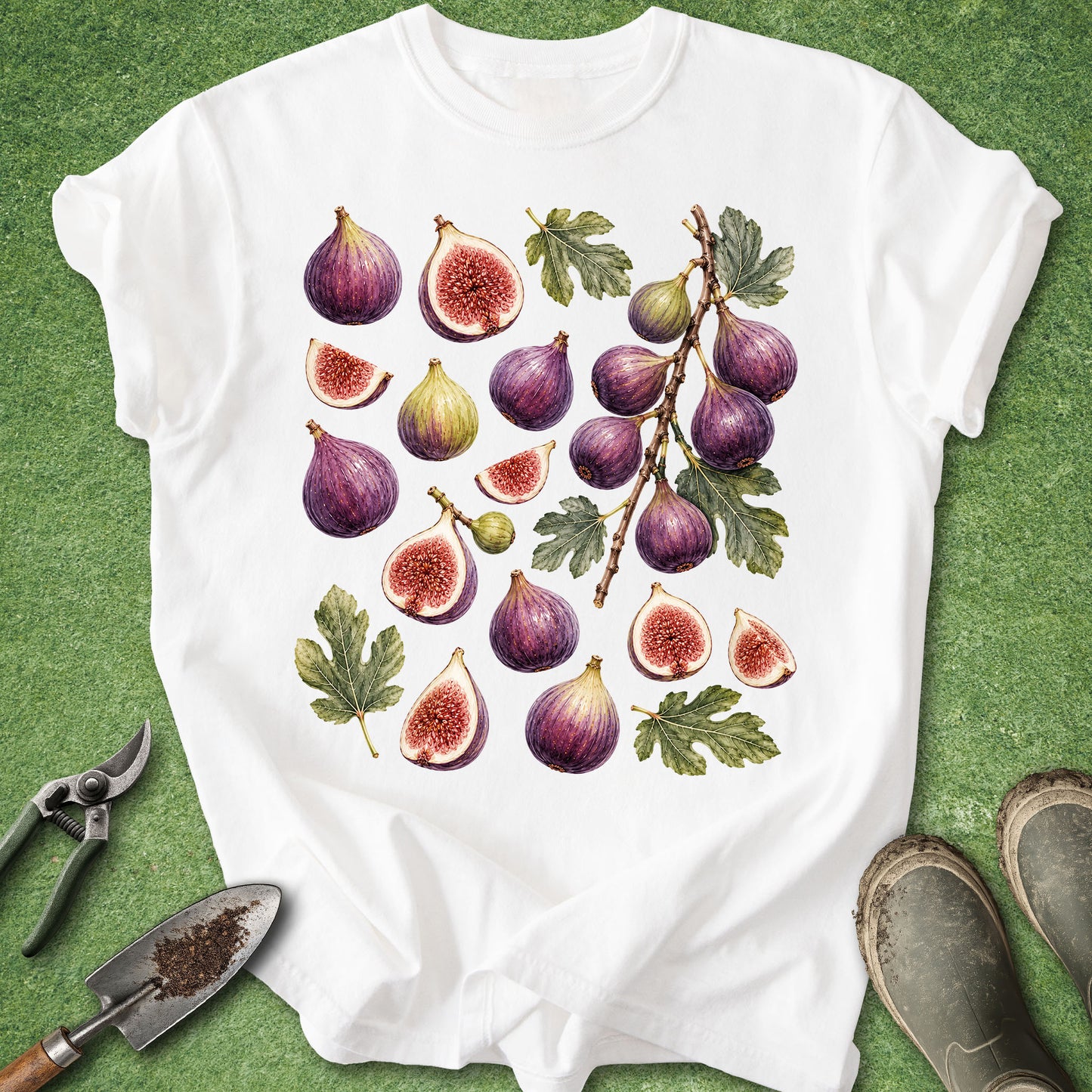 A white short-sleeve T-shirt laid flat on a green grass-like surface, featuring a large illustrated print of purple figs with green leaves. Gardening props including pruning shears, a small hand trowel, and muddy boots are placed around the shirt.