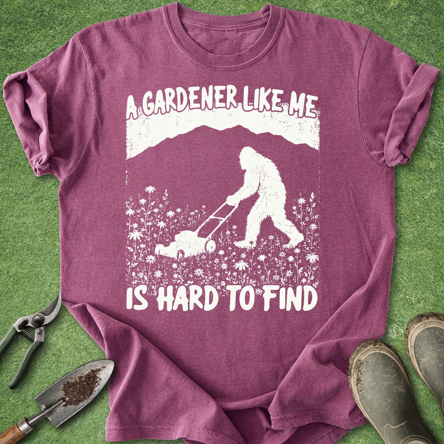 Purple t-shirt with a graphic of a person and Bigfoot gardening, text 'A gardener like me is hard to find', on a grass background.