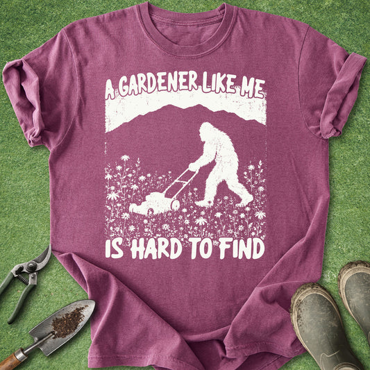 Purple t-shirt with a graphic of a person and Bigfoot gardening, text 'A gardener like me is hard to find', on a grass background.