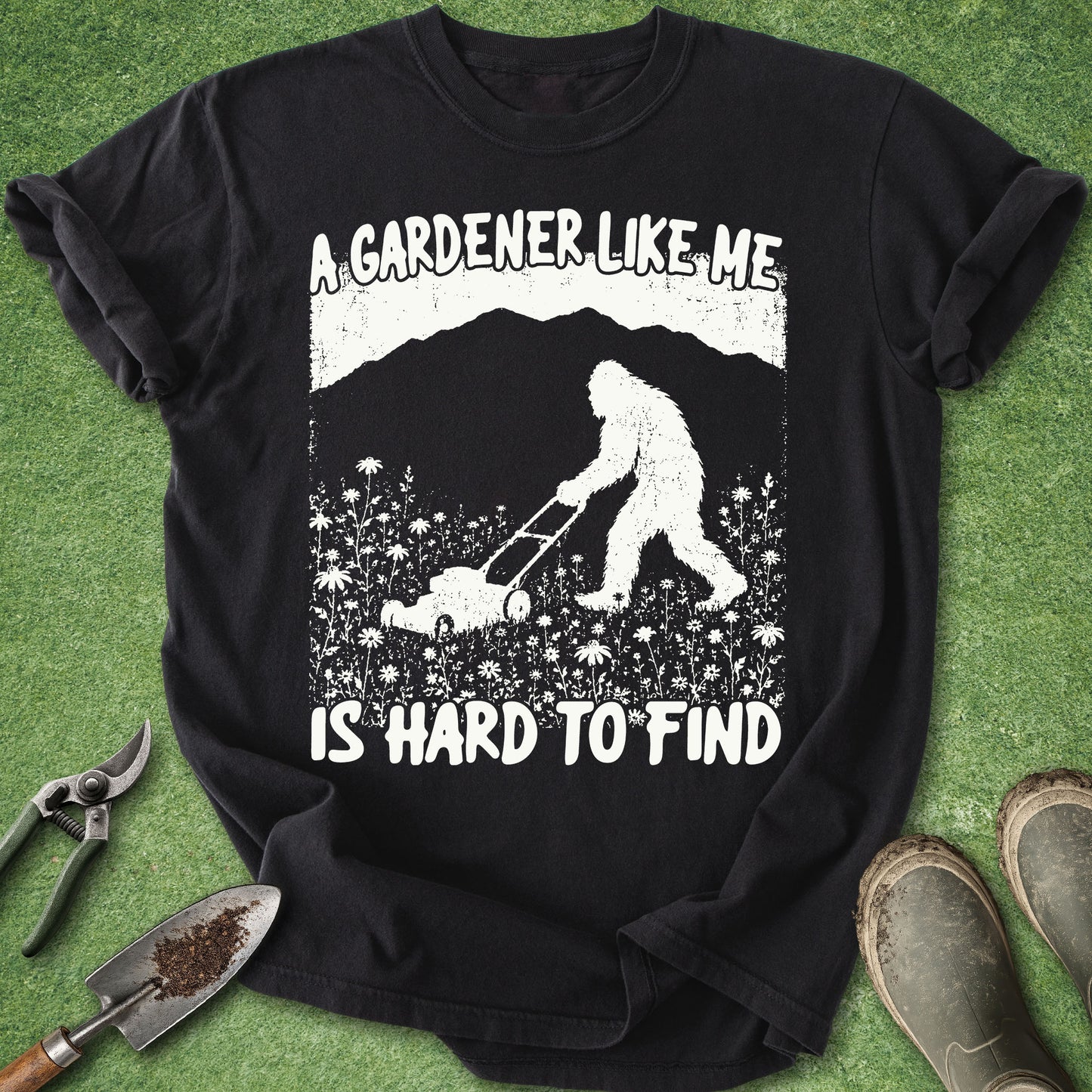 Black t-shirt with a graphic of a person and Bigfoot gardening, text 'A gardener like me is hard to find', on a grass background with gardening tools.