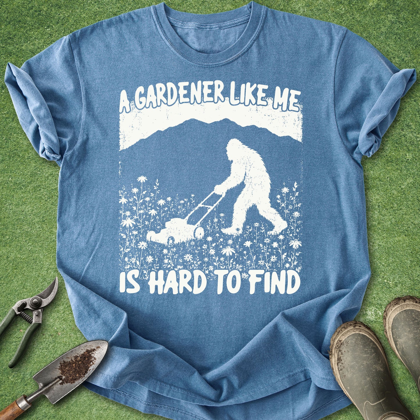 Blue t-shirt with a graphic of a person and Bigfoot gardening, text 'A gardener like me is hard to find', on a grass background.