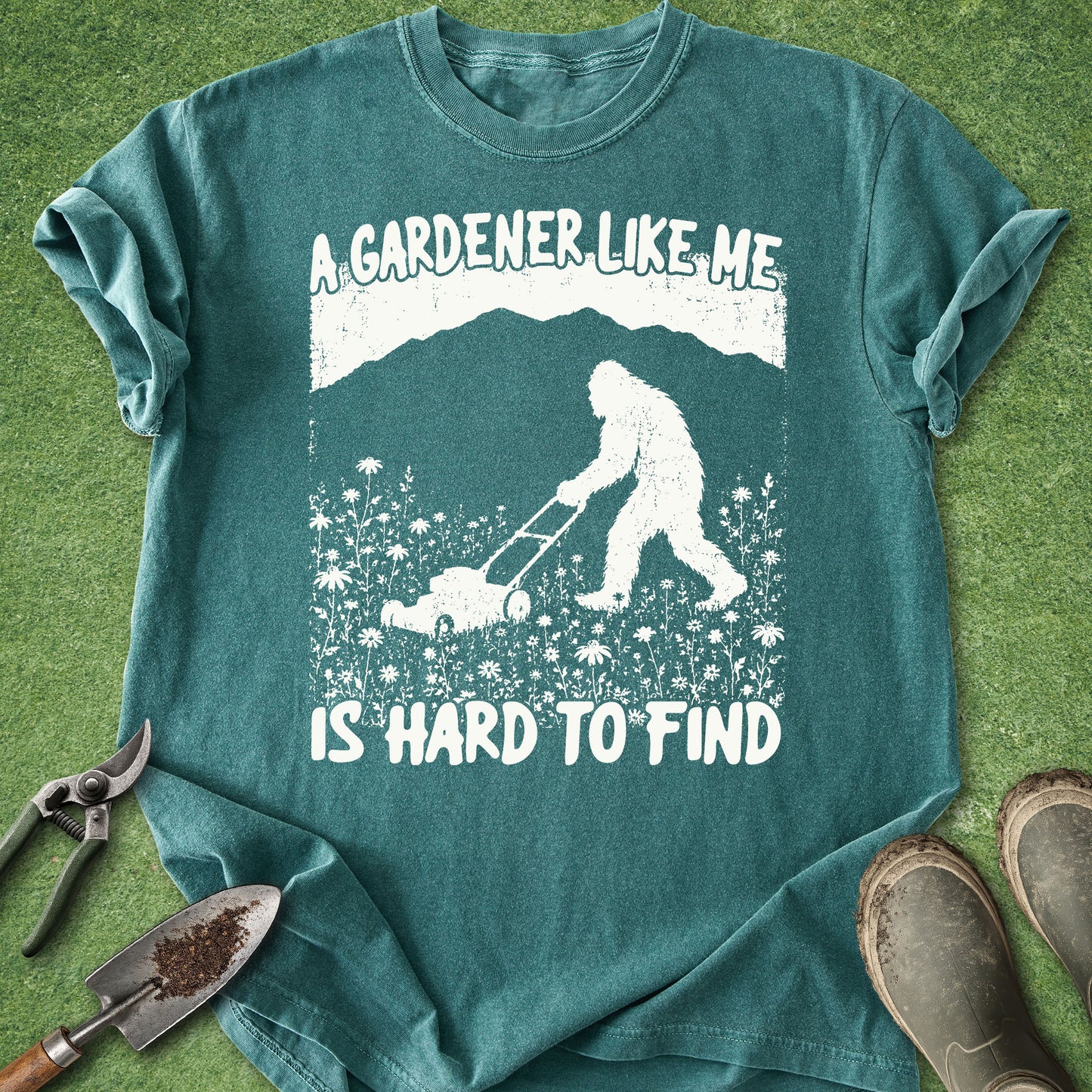 Teal t-shirt with a graphic of a person mowing grass and text 'A gardener like me is hard to find' on a grass background.