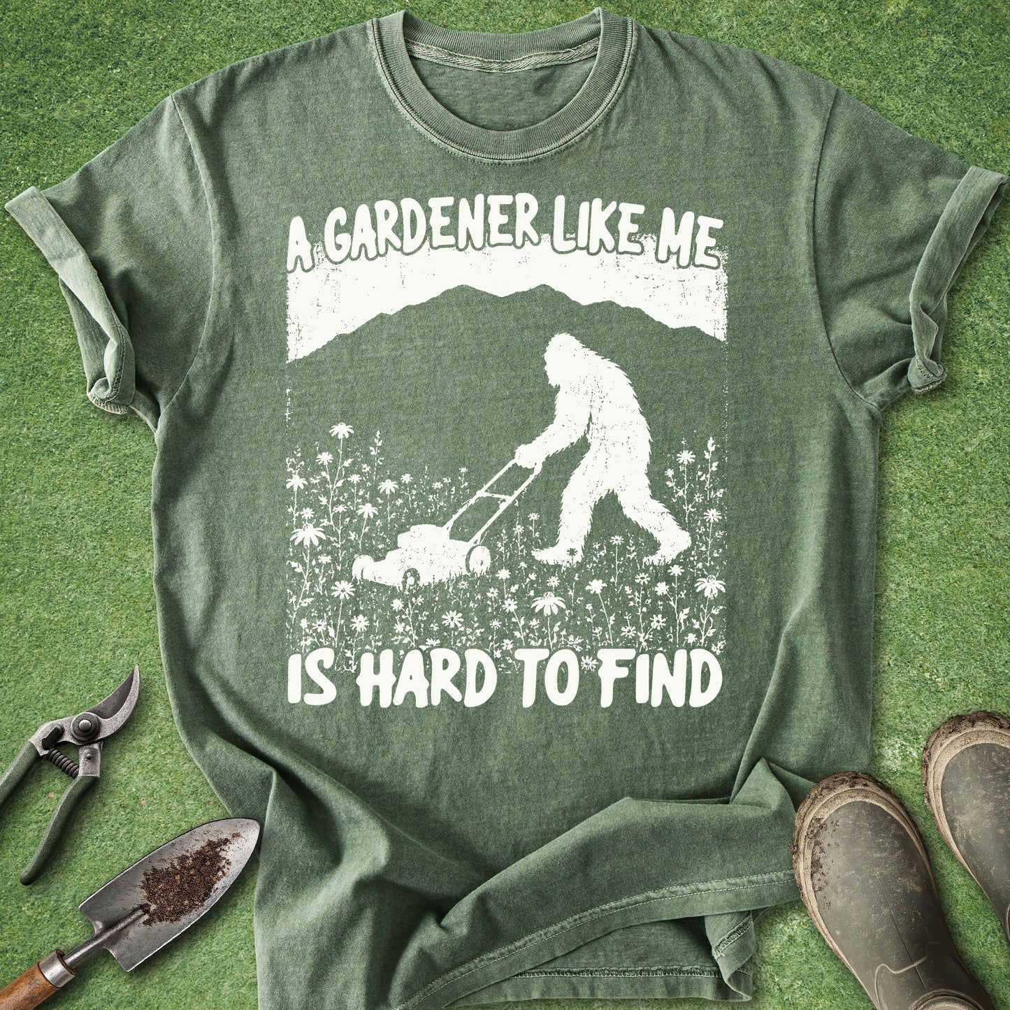 Green t-shirt with a graphic of a person and Bigfoot mowing grass, text 'A gardener like me is hard to find', on a grass background.