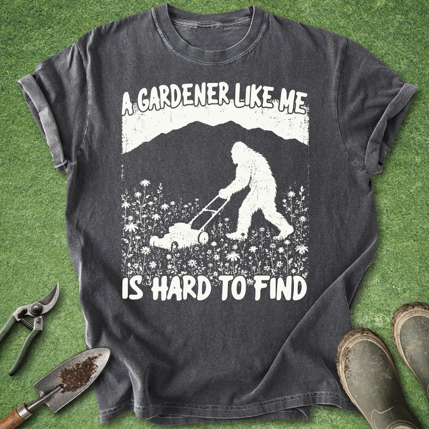 Gray t-shirt with a graphic of a person and Bigfoot mowing grass, text 'A gardener like me is hard to find', on a green grass background.
