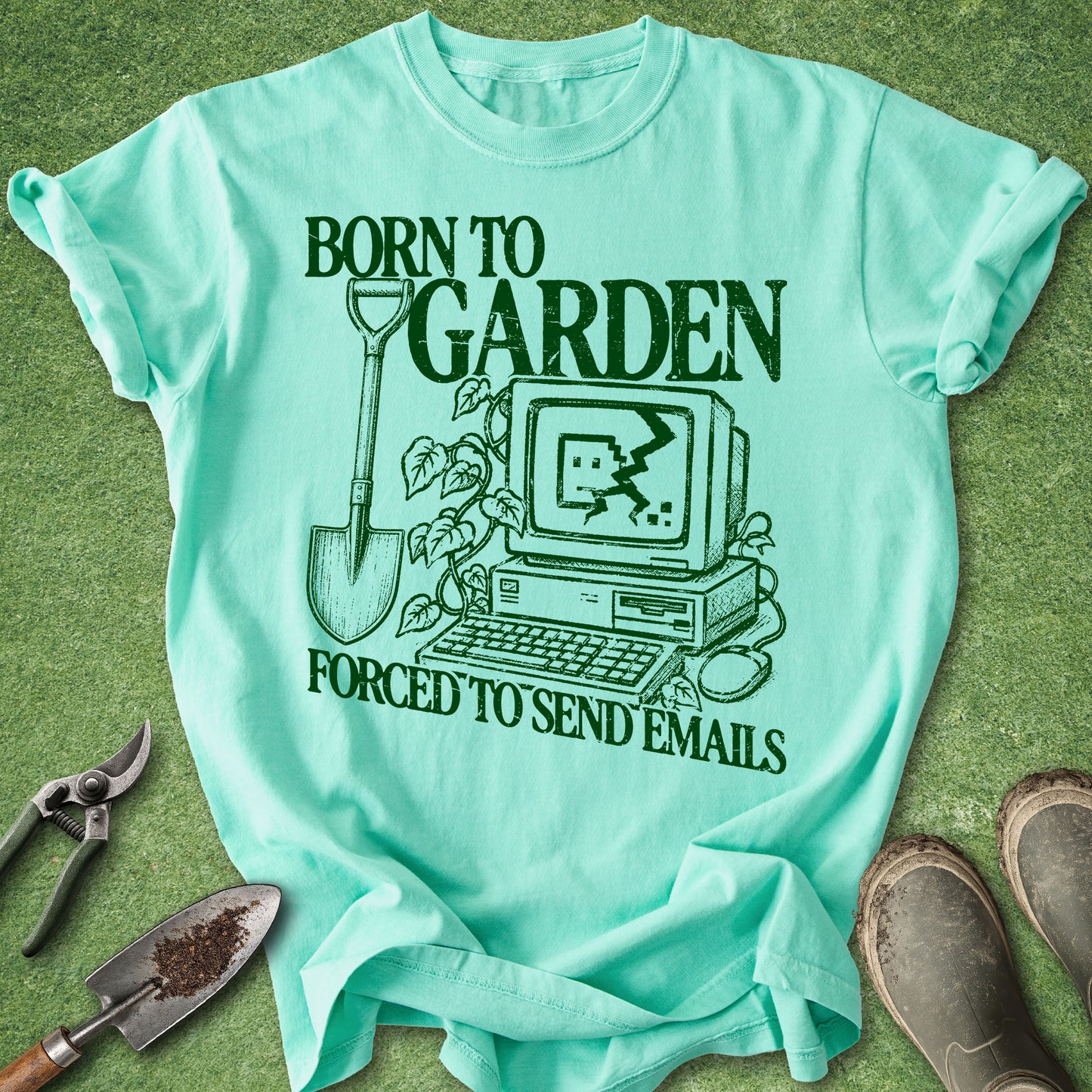 A mint green graphic t-shirt laid flat on a green grass-like surface, surrounded by gardening props including pruning shears, a trowel, and work boots. The shirt features a dark green graphic of a computer, shovel, and plants with the text 'BORN TO GARDEN FORCED TO SEND EMAILS'.
